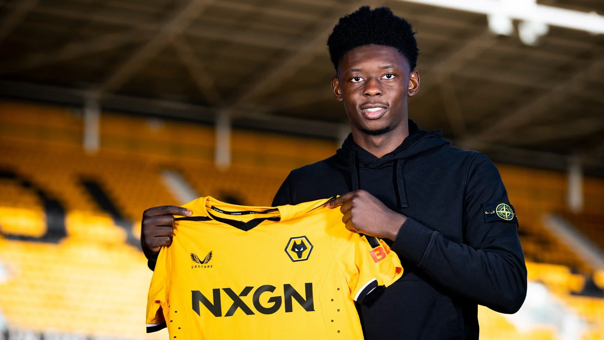 Young man holding a yellow Wolverhampton Wanderers football jersey inside a stadium.