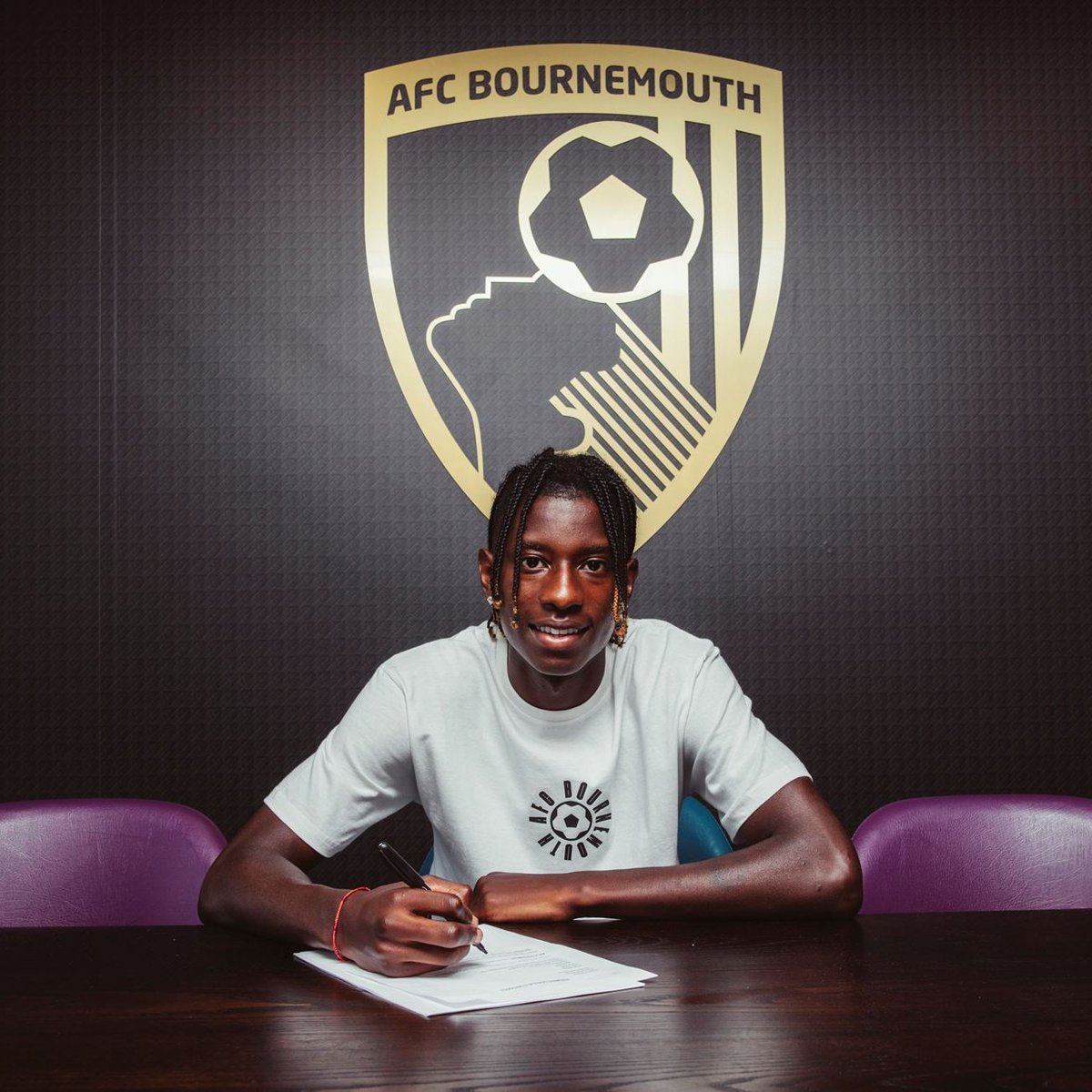 Young man sitting at a table signing documents at AFC Bournemouth signing event, with team logo on the wall behind him.