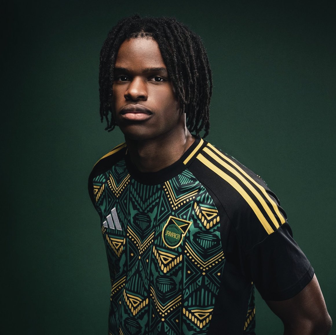 Portrait of a young man with dark skin, dreadlocks, wearing a black, green, yellow, and gold Jamaican football jersey with Adidas logo on the chest and a club emblem reading Jamaica.