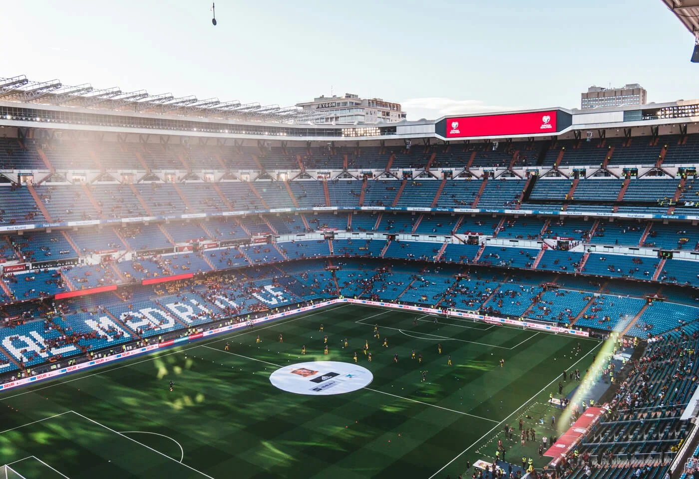 A nearly empty soccer stadium with a green field, seating areas, video screen, and players warming up.