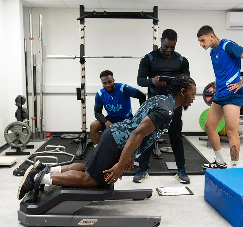 A group of men in a gym, with one man using a training machine, all wearing football academy practice gear.