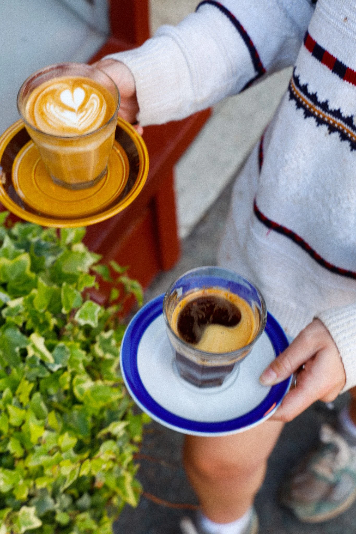 Person holding two small glasses of espresso and latte with decorative foam, standing outdoors near greenery.