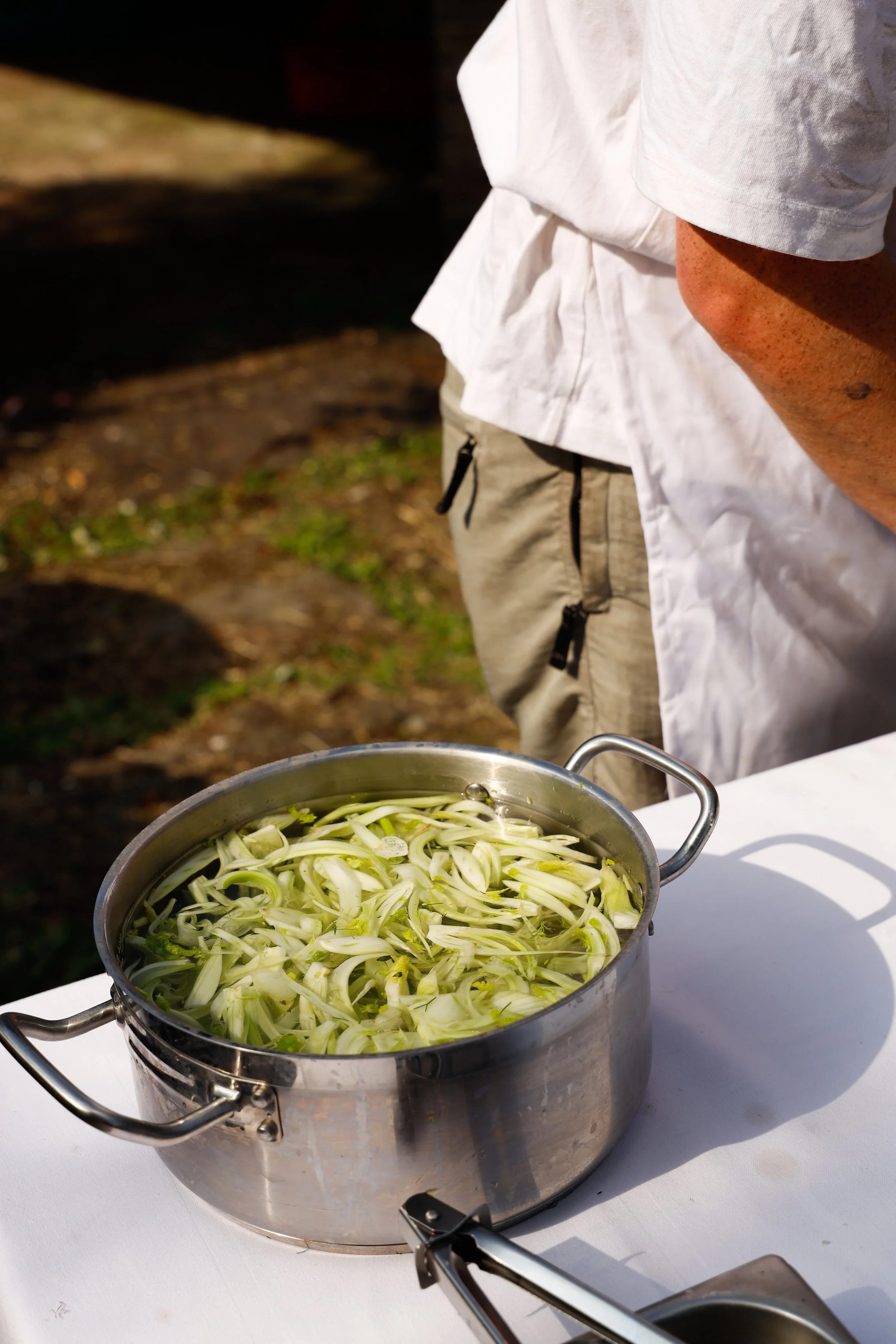 A metal pot filled with sliced fennel on a table, with a person wearing a white shirt standing nearby outdoors.