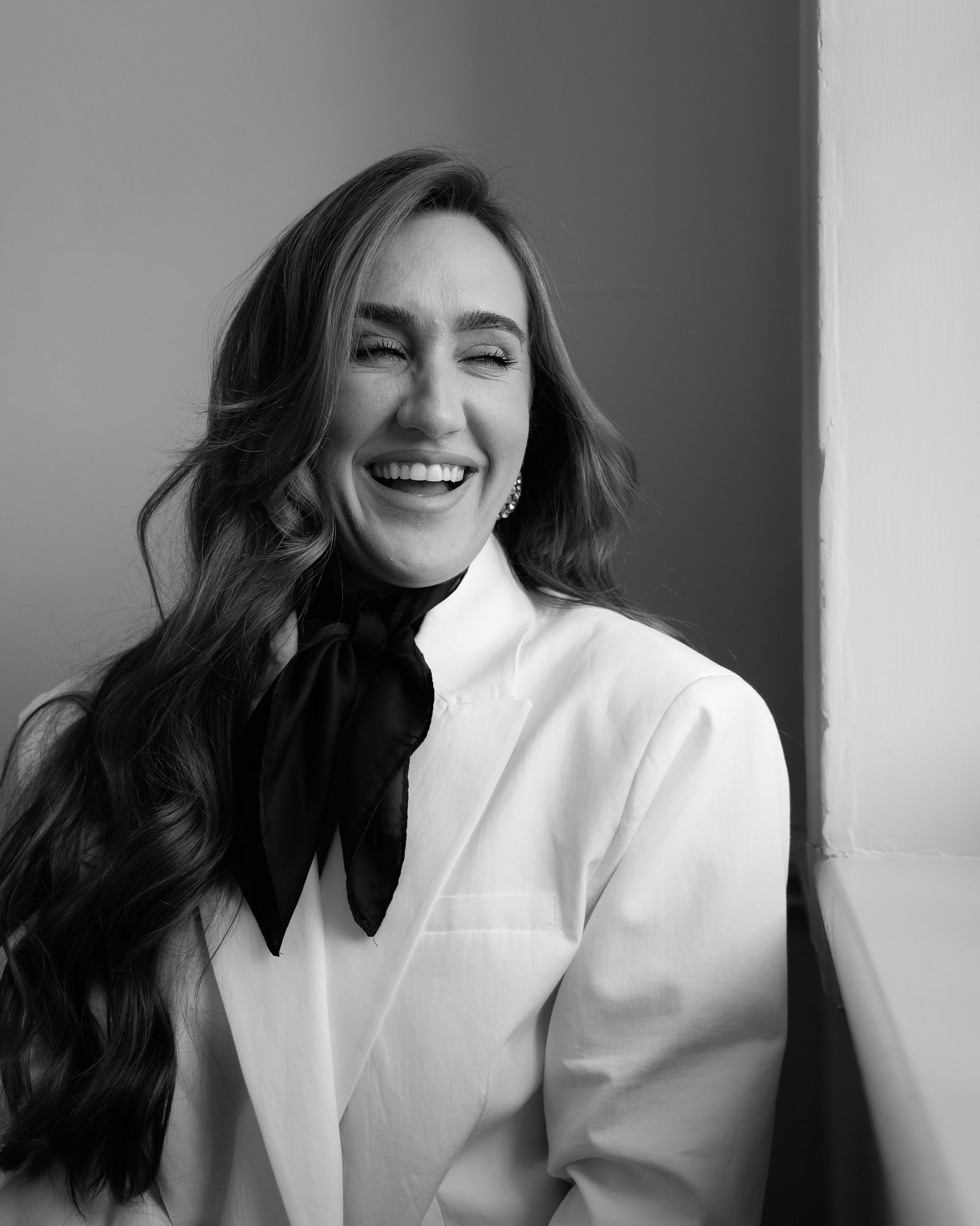 Black and white photo of a woman with long wavy hair, smiling and looking to her right, wearing a white blazer and a dark scarf tied around her neck, leaning against a wall.