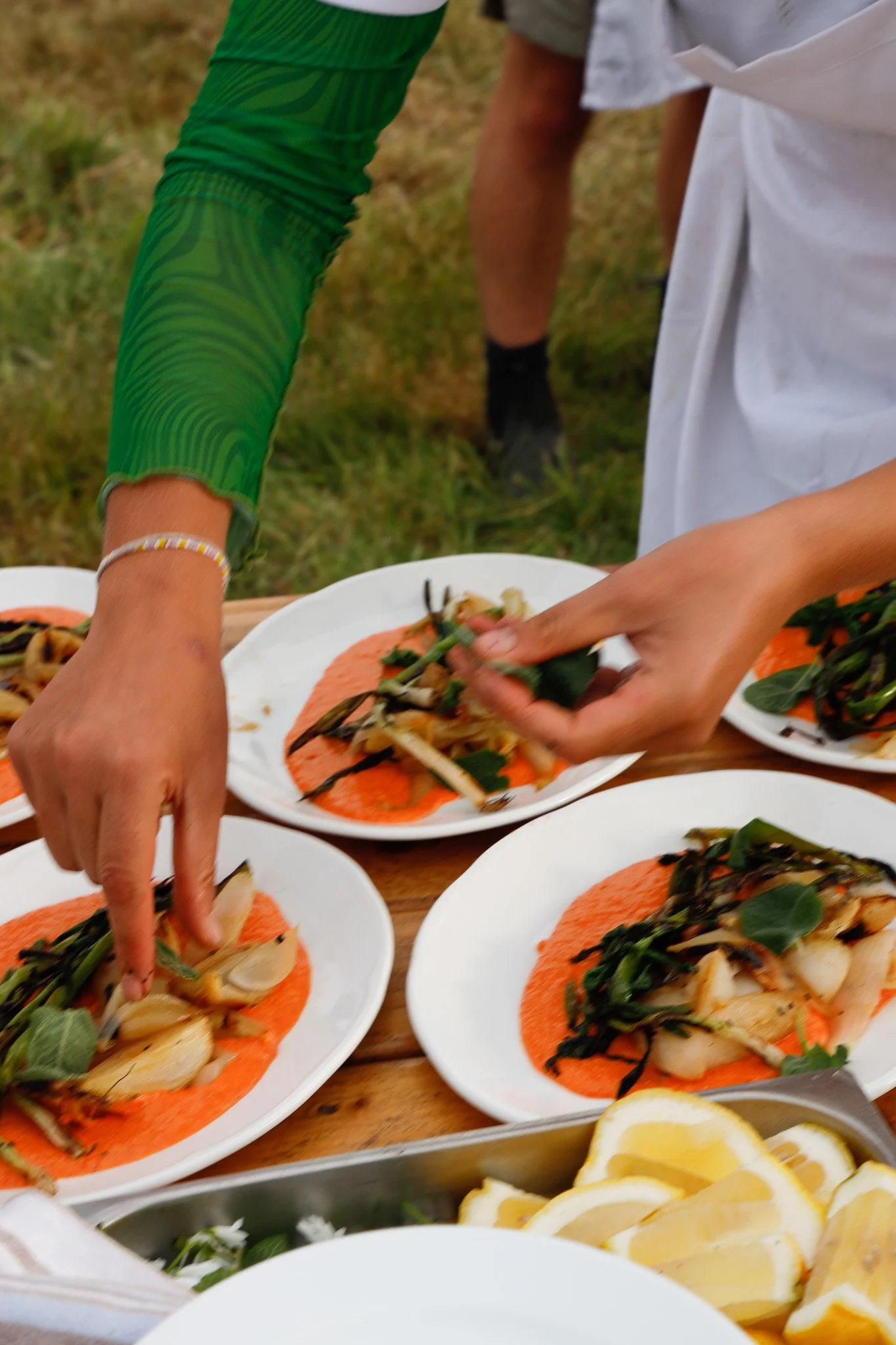Close-up of several plates of roasted scallops and vegetables on a wooden table, with hands of people serving the food, outdoors during daytime.