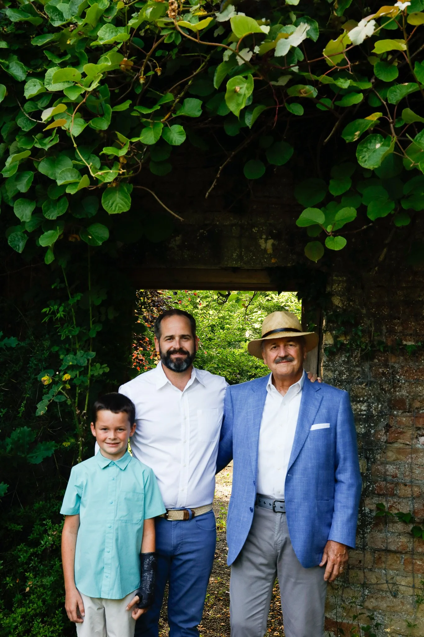 Three males standing together in front of an archway covered with green leaves, smiling at the camera. The young boy is on the left, the man with a beard and white shirt is in the middle, and the elderly man with a hat and blue blazer is on the right