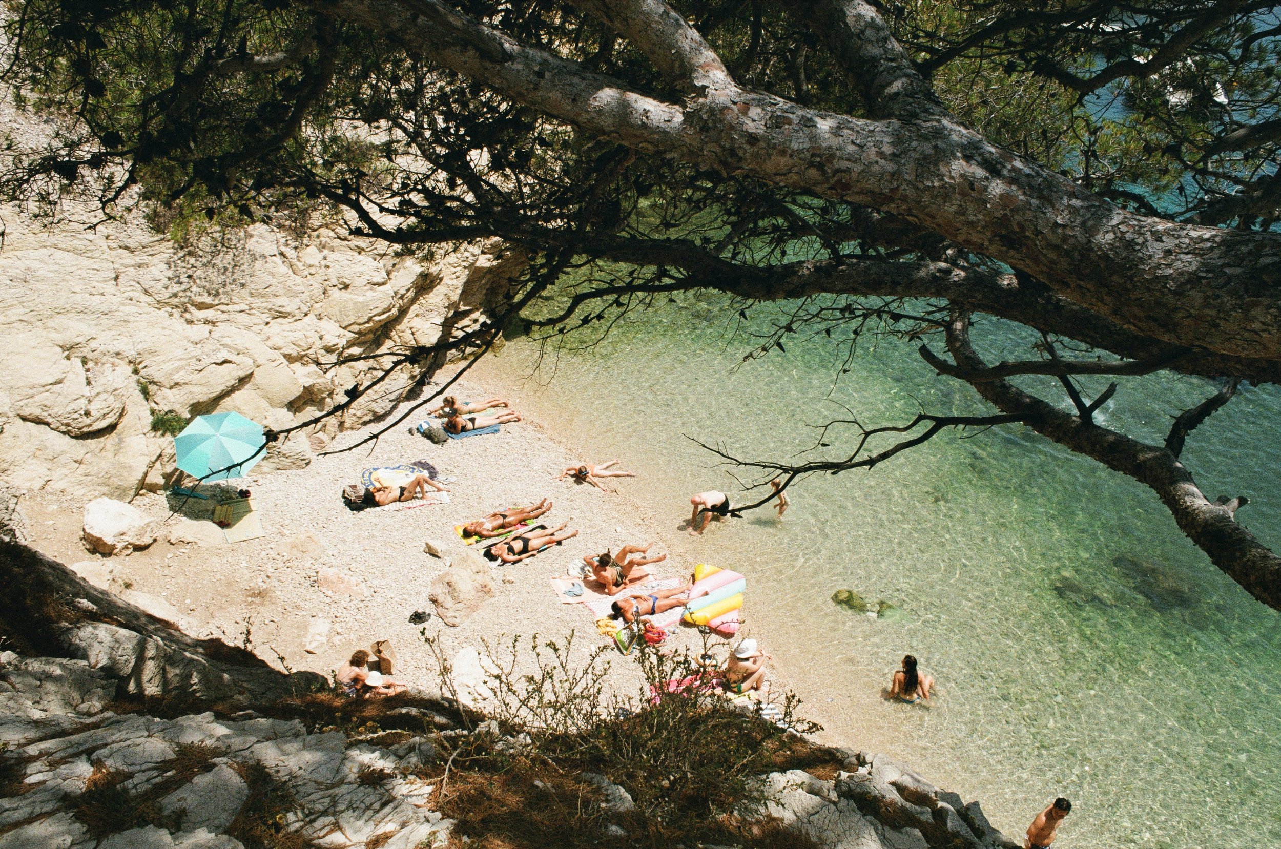 People sunbathing and swimming on a rocky beach shaded by trees, with clear water and a small float.