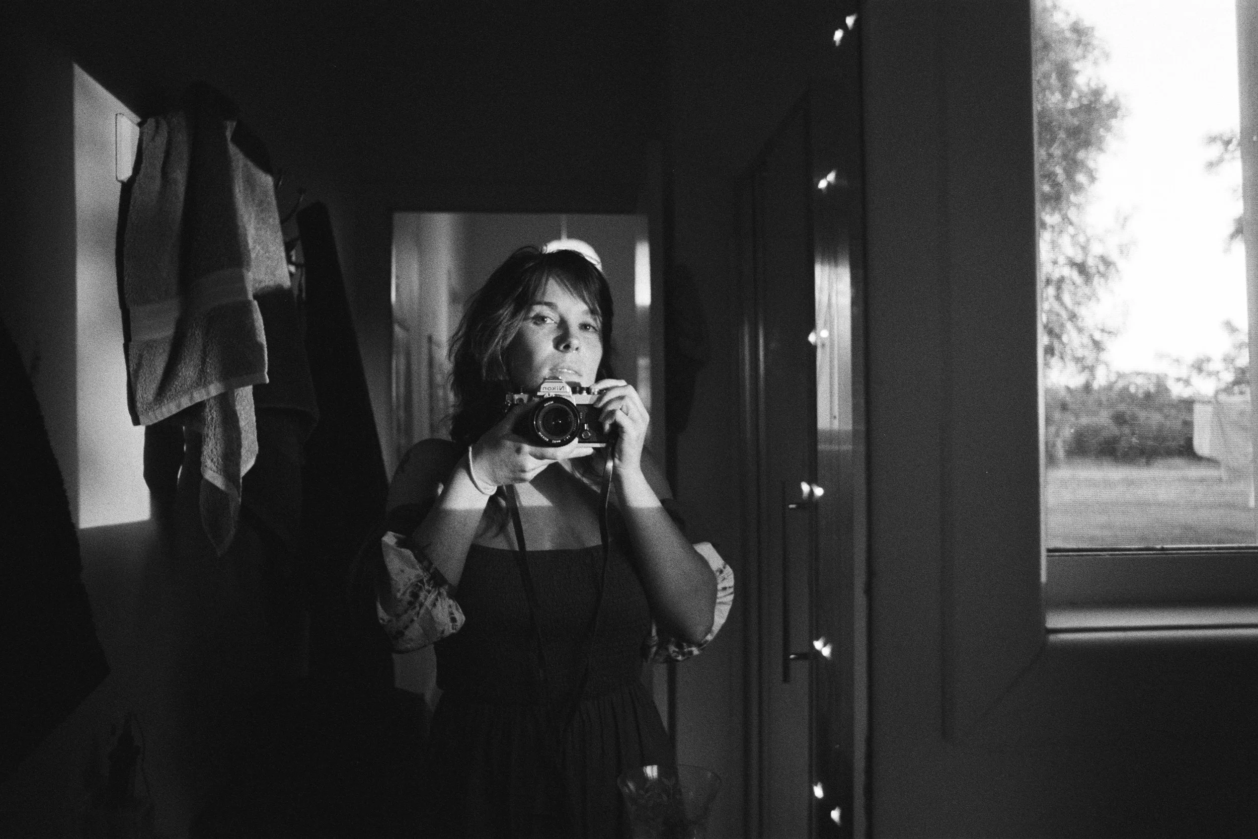 A woman taking a selfie in a mirror with a camera indoors next to a window showing a yard and trees outside.
