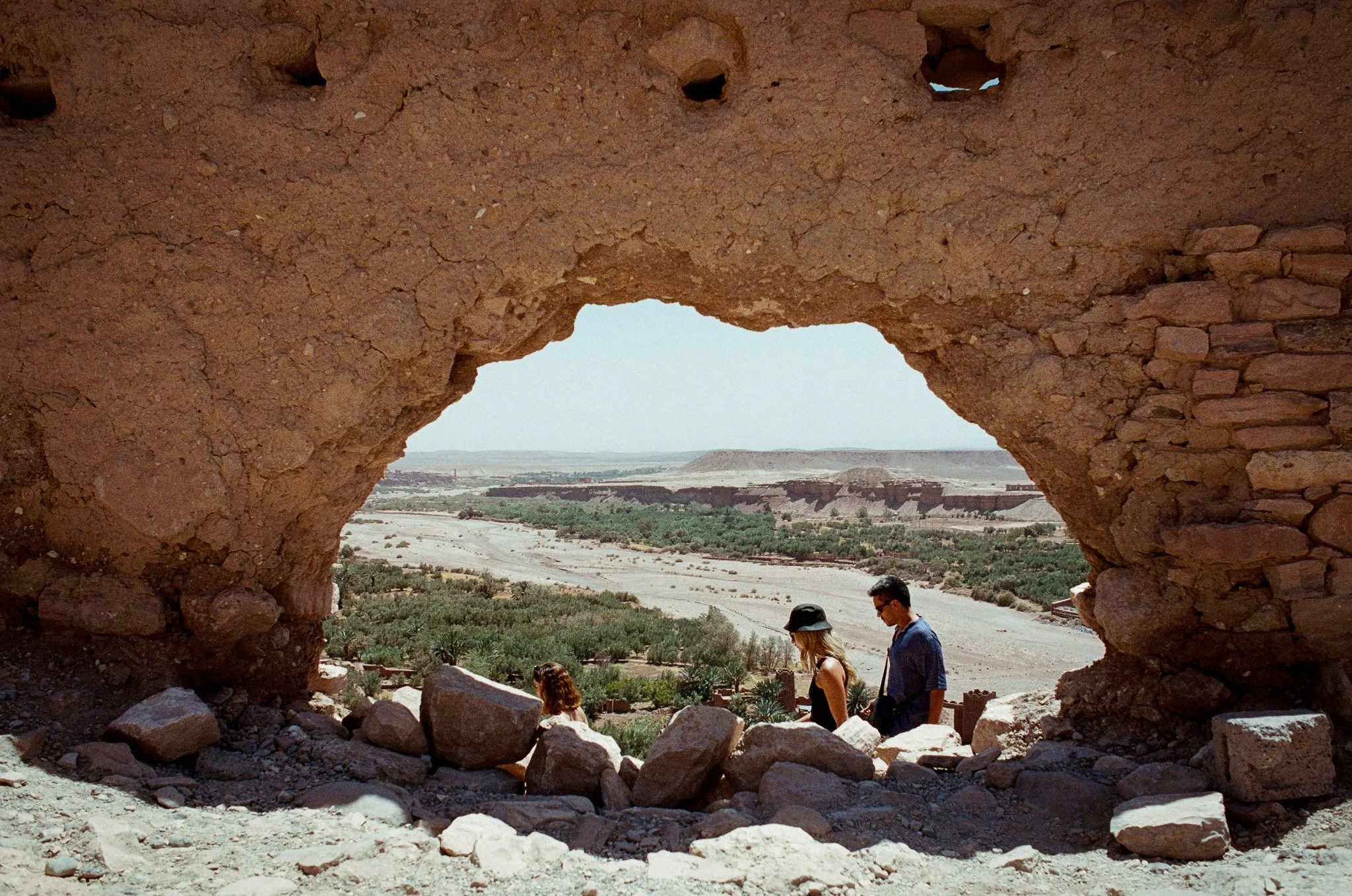 Three people walking through a desert landscape seen through a large, irregularly shaped opening in a weathered, earthen wall, with a few holes at the top and scattered rocks at the bottom.