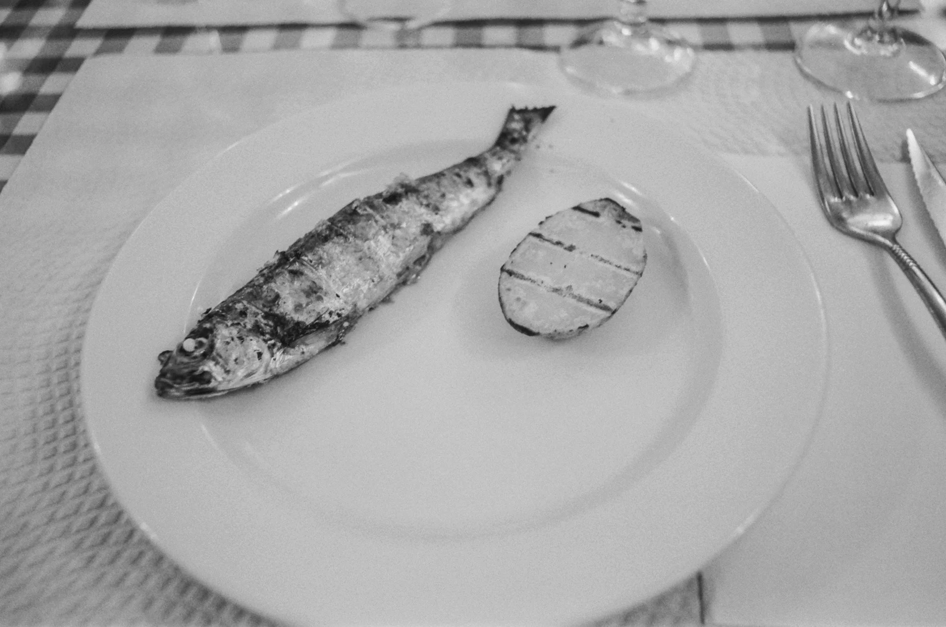 A whole grilled fish on a white plate with a side of grilled lemon half, set on a table with a fork and knife to the right.