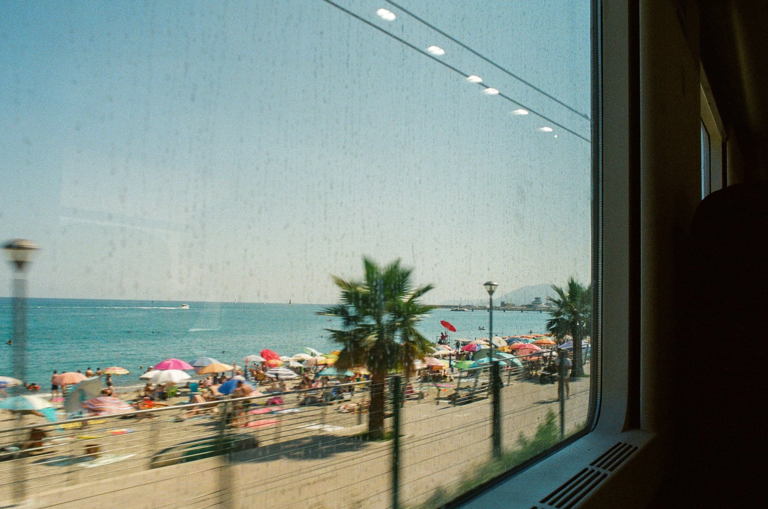 Beach scene seen through a train window, with colorful umbrellas, palm trees, and people on the sandy shore, and the ocean with boats in the background.