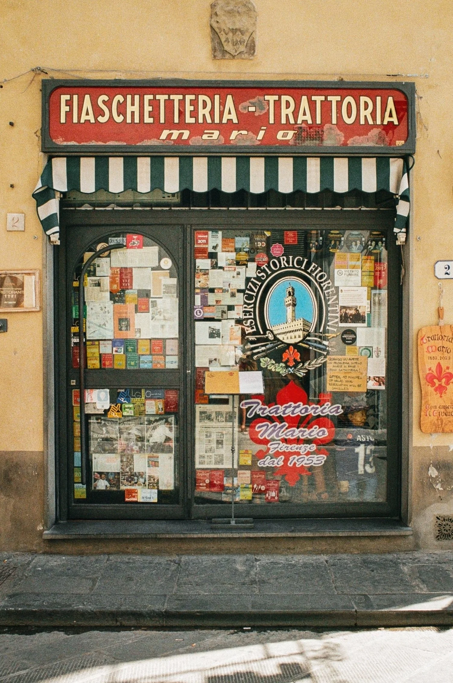 The storefront of Trattoria Mario, an Italian restaurant in Florence with a red and green striped awning, handwritten signs, and a window display filled with notices and decorations.