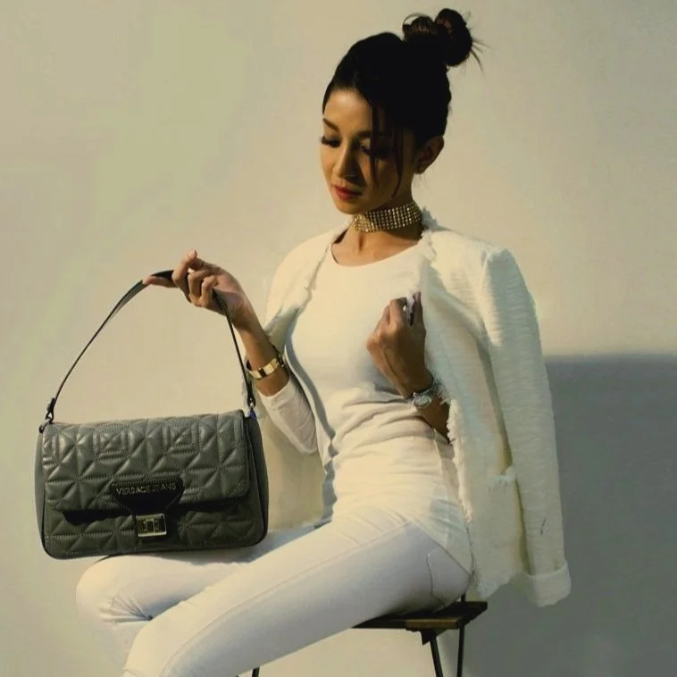 Femme élégante assise sur une chaise blanche, portant un ensemble blanc avec un blazer et un jean, avec un sac à main gris en quilté, portant un collier ras-de-cou doré et un bracelet, avec ses cheveux attachés en haut.