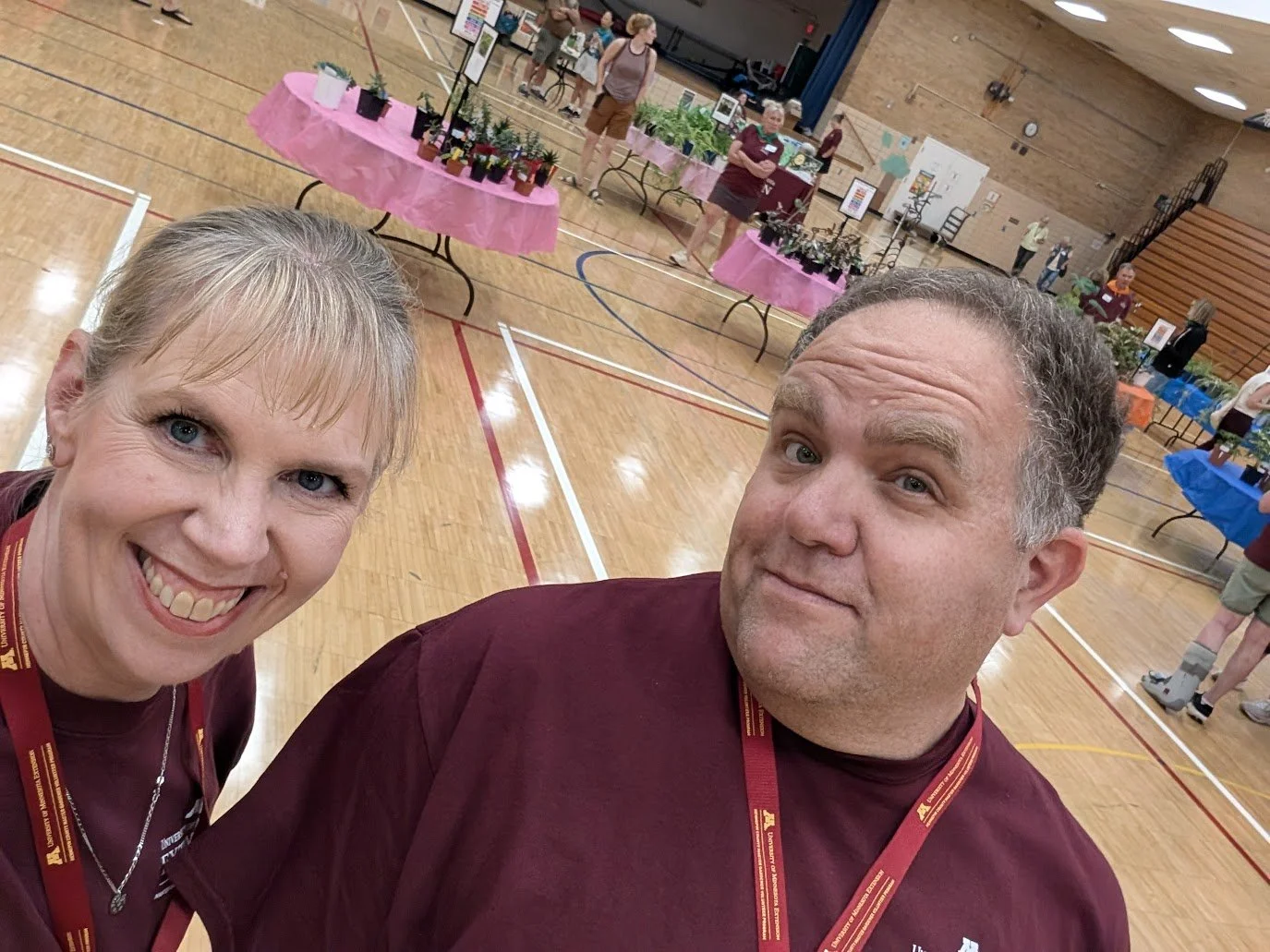 Two people taking a selfie at an indoor event with tables of potted plants and people browsing in the background.
