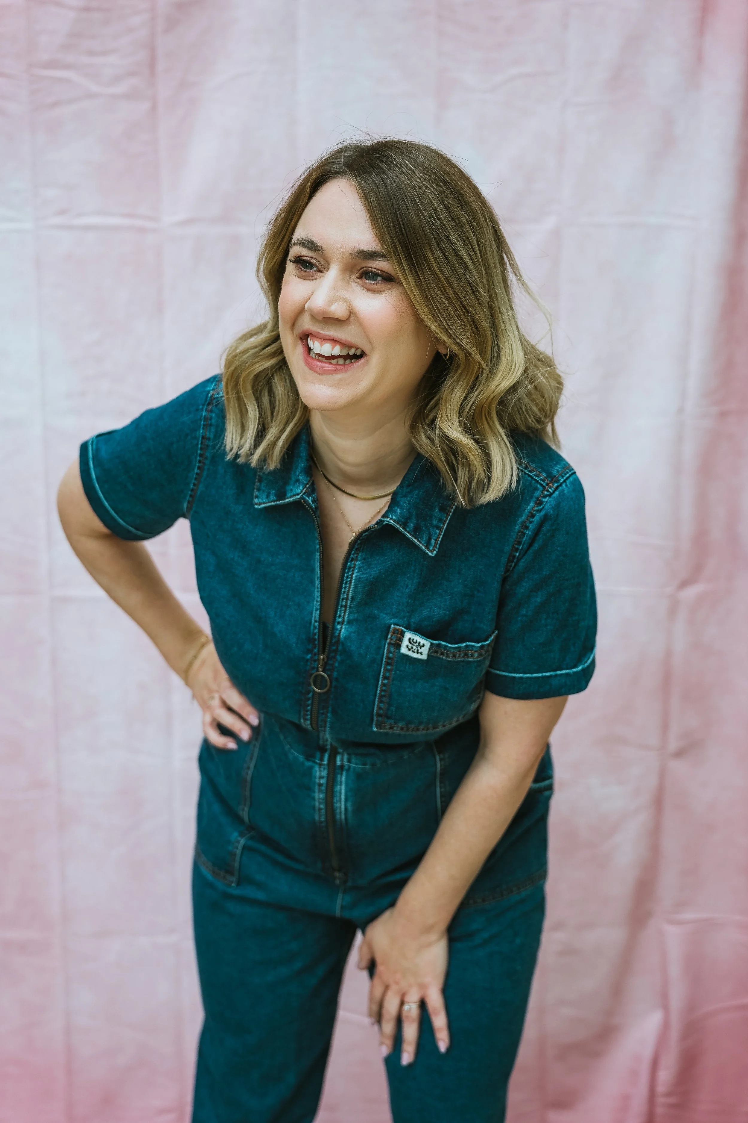 Fiona Quiggin; a woman with shoulder-length blonde hair, wearing a blue denim jumpsuit, laughing with her hand on her hip and leaning forward, against a pink fabric background.