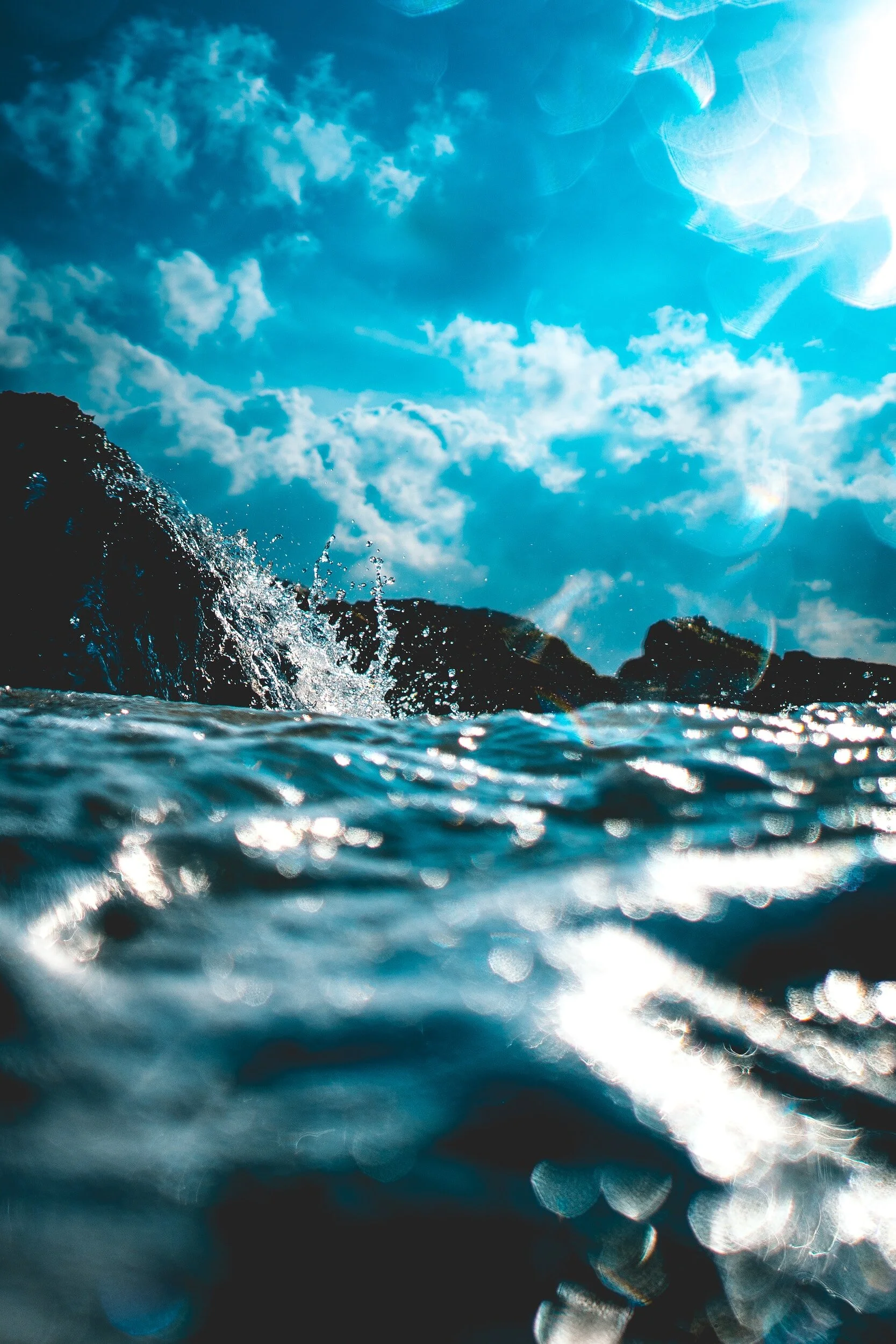 Close-up of ocean water with rocks in the distance and blue sky with clouds, sunlight creating glimmers on the water surface.