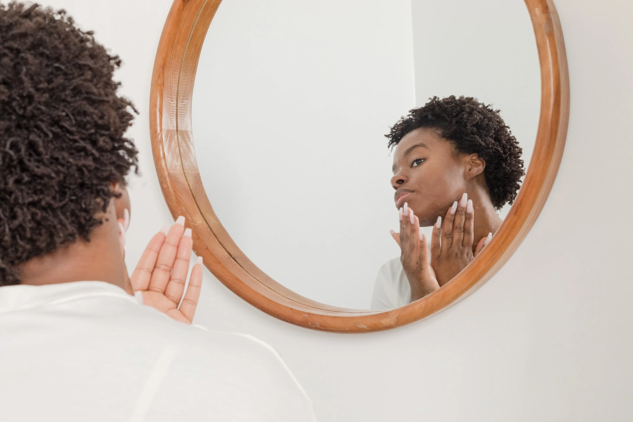 A woman with short, curly hair looking at her reflection in a round wooden-framed mirror, touching her face with both hands.