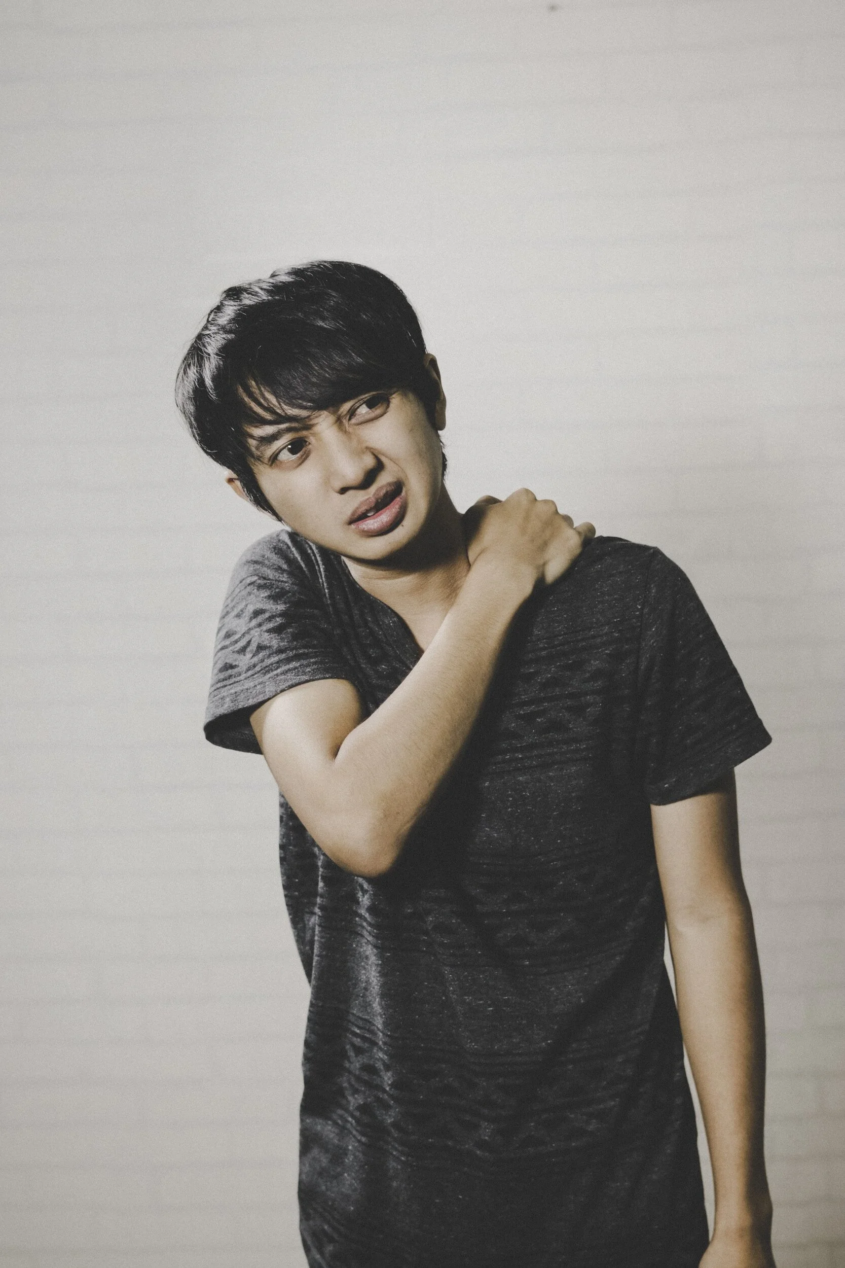 Young man wearing a dark t-shirt, holding his shoulder, with a pained or uncomfortable expression.