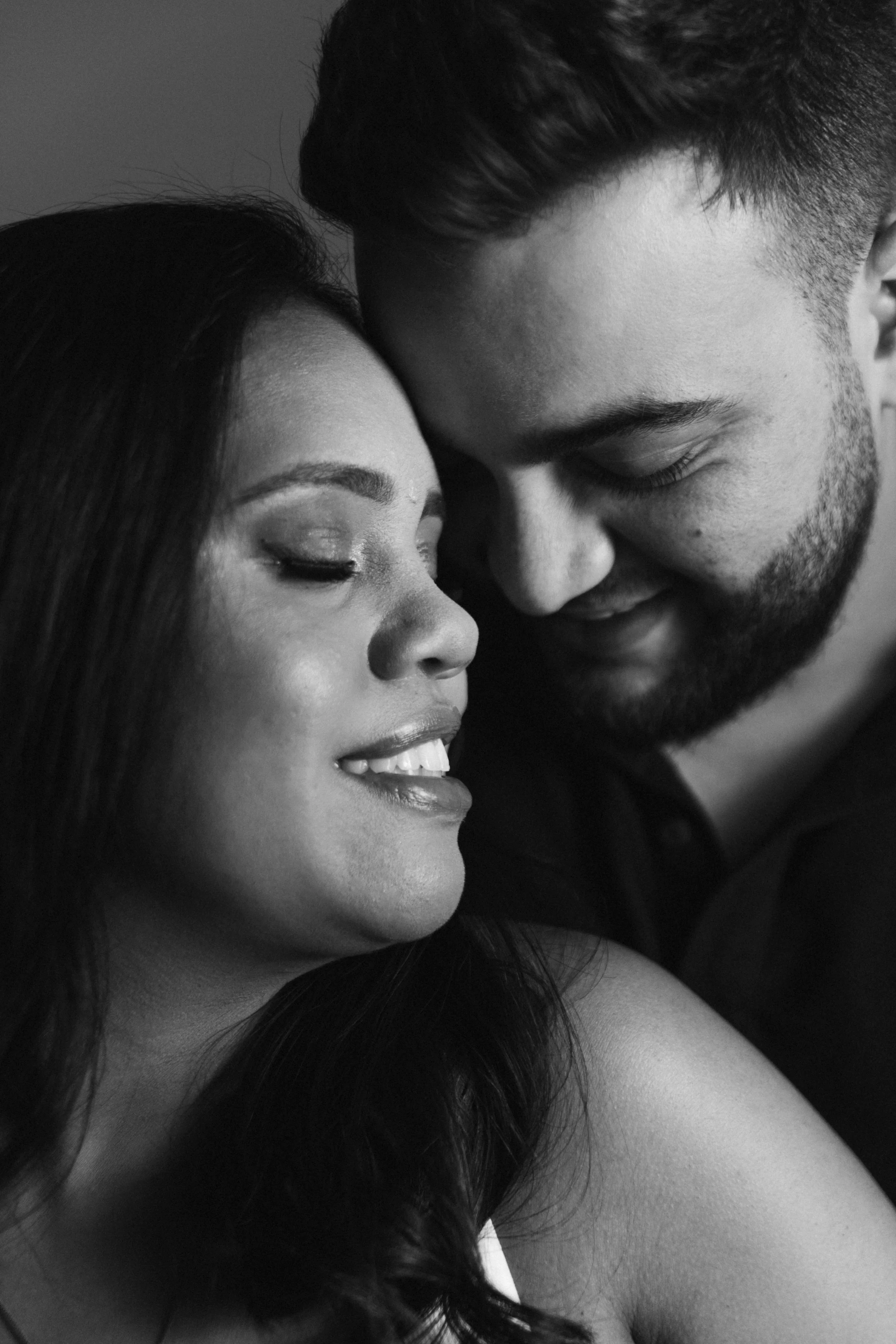 Close-up black and white photo of a couple with their foreheads and noses touching, smiling gently with eyes closed.