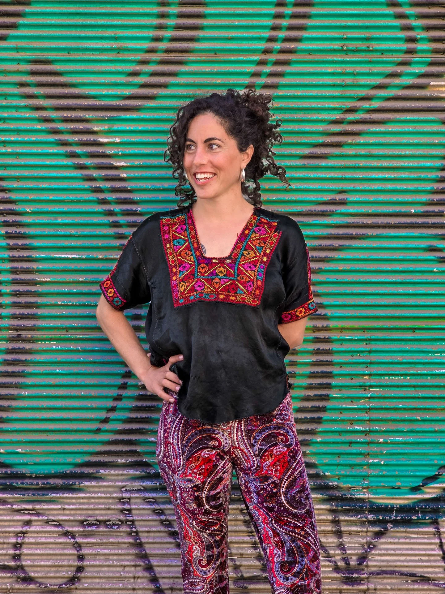 A woman with curly dark hair, wearing a black embroidered top and patterned red and purple pants, standing in front of a colorful graffiti wall.