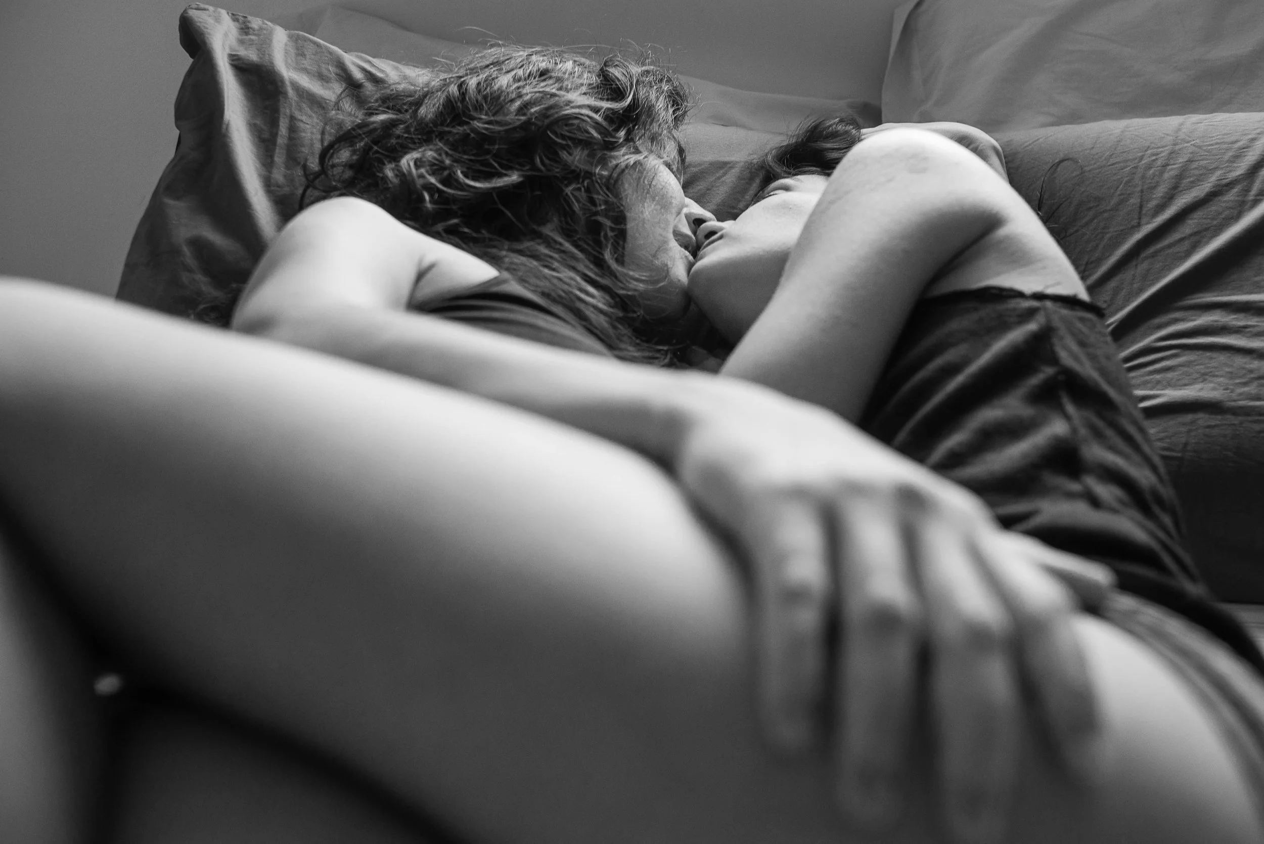 A black and white photograph of two women lying closely together on a bed, embracing each other affectionately.