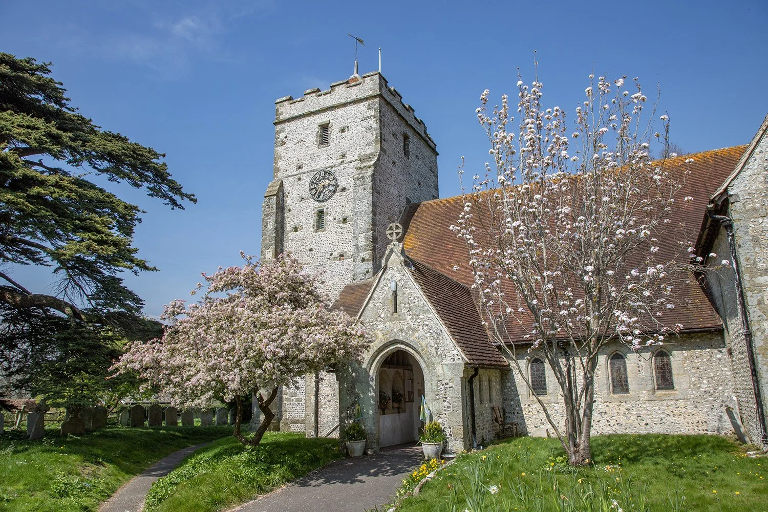 Historic stone church with tower and clock, surrounded by springtime blooming trees and greenery.