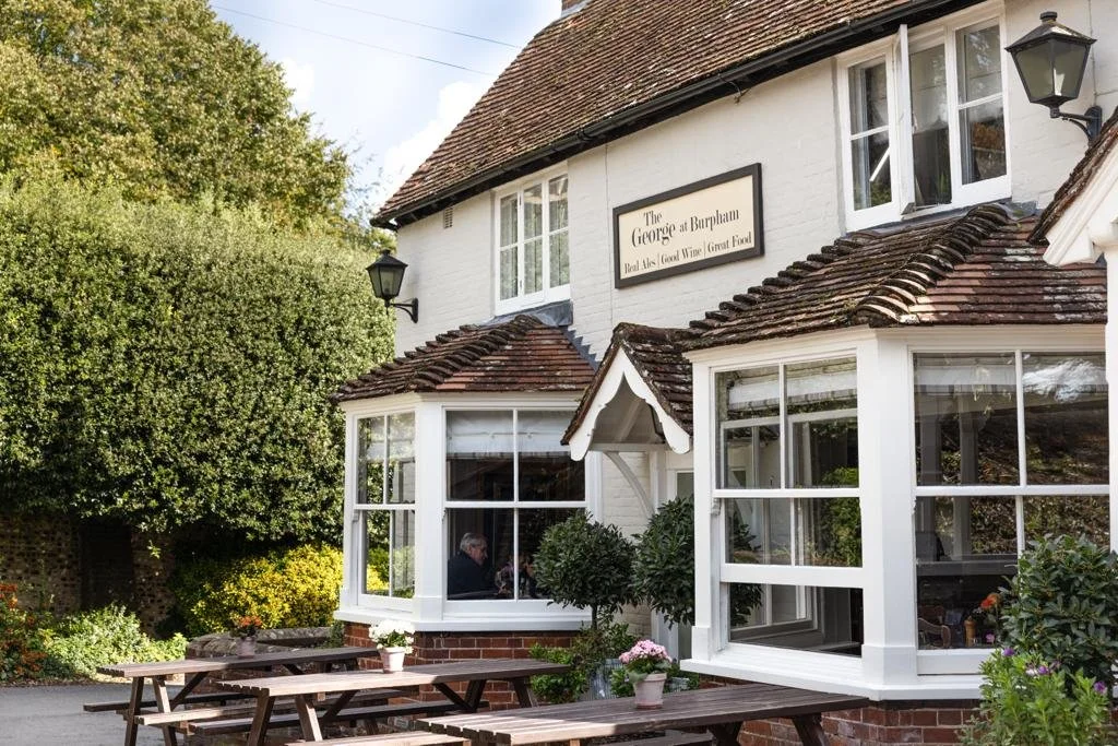 Exterior of The George at Burpham pub, featuring white brick walls, a sign, large windows, and outdoor seating with picnic tables.