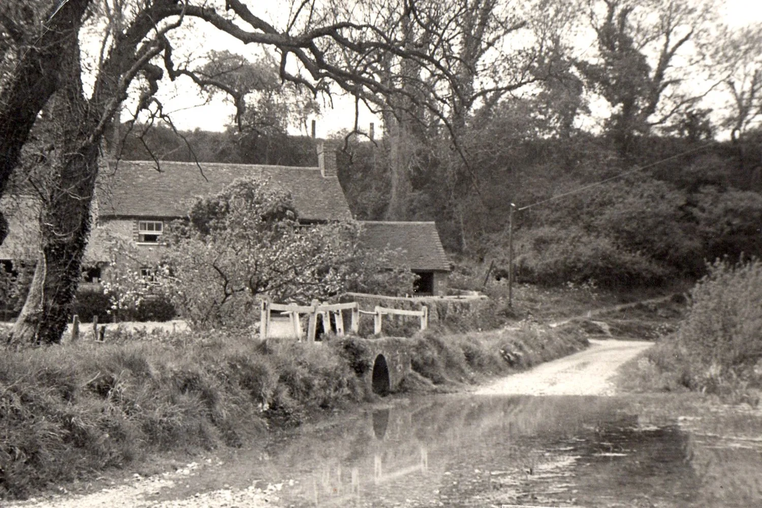 Vintage photograph of a rural landscape featuring a small country house with a thatched roof, surrounded by trees and foliage. A narrow dirt road runs beside the house, crossing a shallow stream.