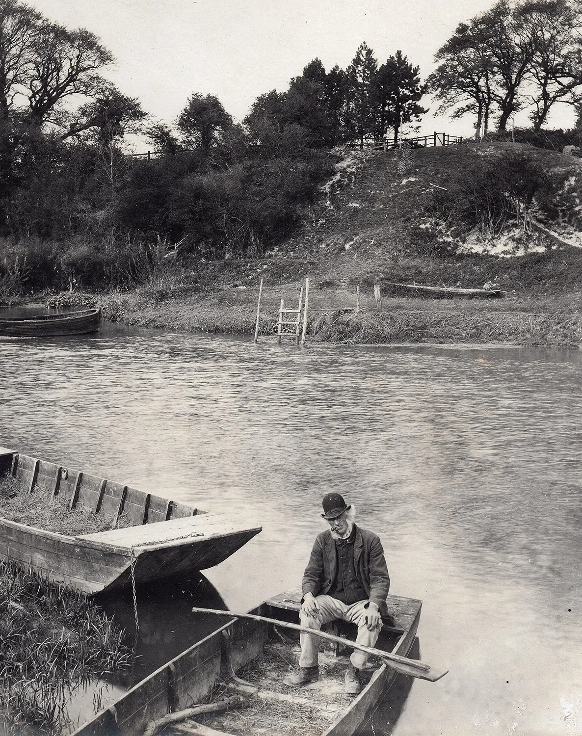 Vintage photo of a man in a small wooden boat on a river, with a second empty boat nearby. The riverbank is lined with trees and a grassy hill in the background.