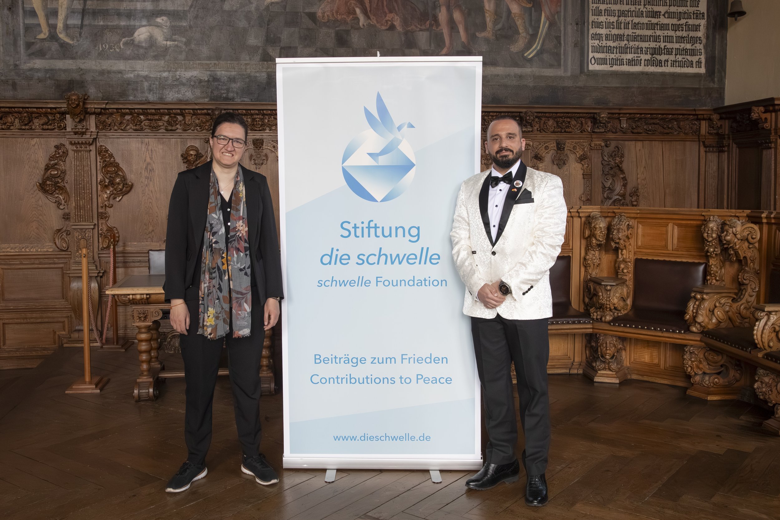 Two individuals dressed in formal attire standing next to a large banner with the logo and text for Stiftung die schwelle, a German foundation. The background features ornate woodwork and historic artwork or murals.