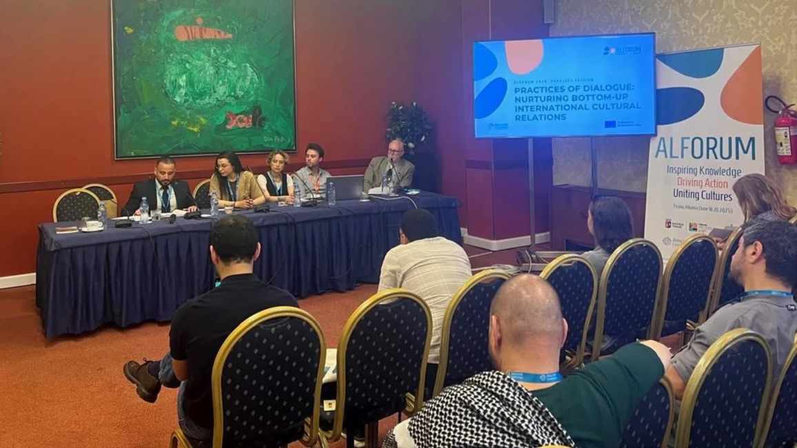 Panel discussion at a conference with a group of five people sitting at a long table facing an audience. A large screen displays a presentation titled 'Practices of Dialogue: Nurturing Bottom-Up International Cultural Relations'. An ALFORUM banner is