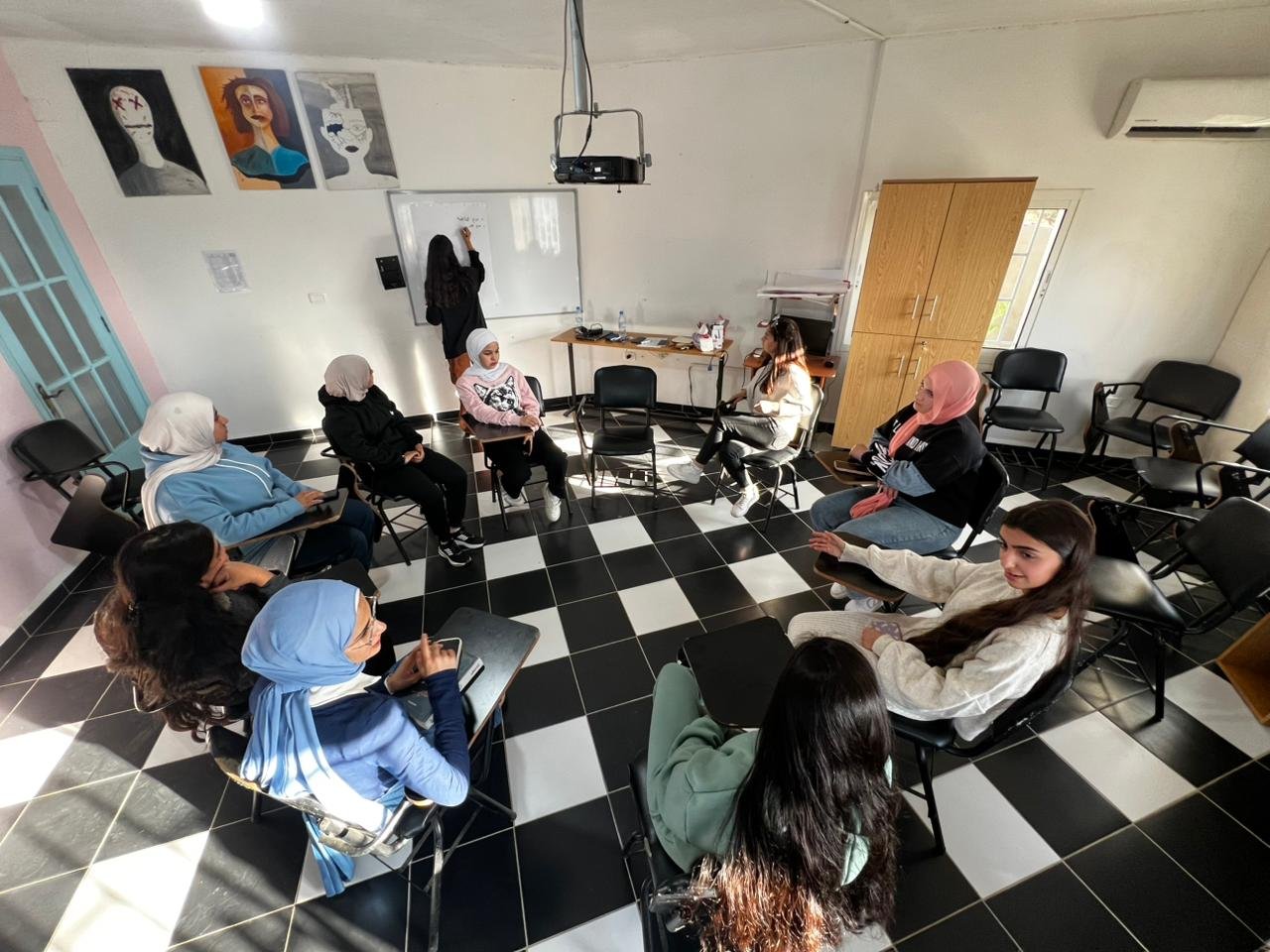 A group of women arranged in a circle in a classroom, some wearing headscarves, participating in a discussion or workshop with a woman at the whiteboard.