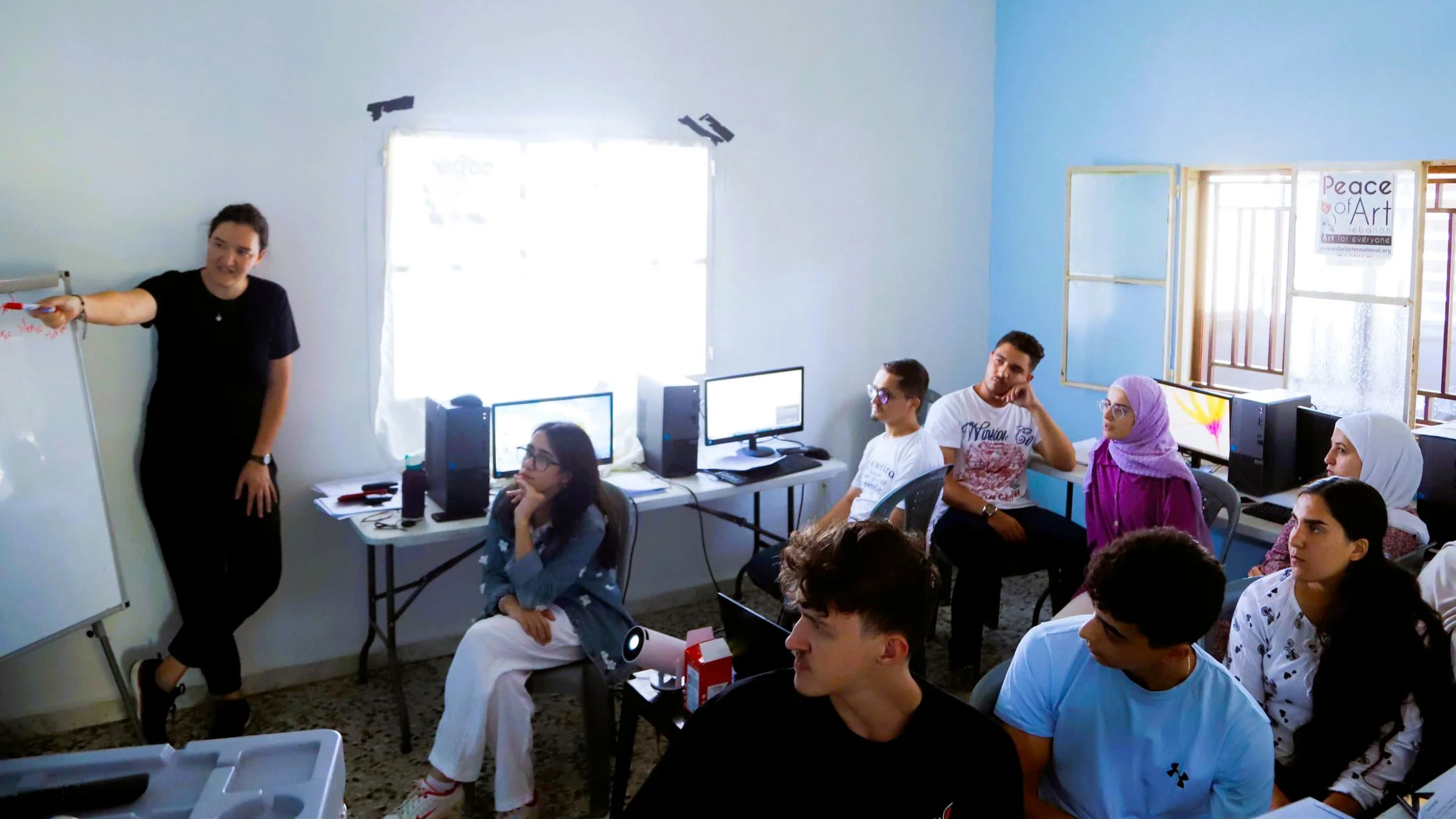 A classroom with students attentively listening to a teacher who is standing near a whiteboard. The room has several computers on desks, and some students are wearing glasses and headscarves. There is a sign on the window that reads "Peace of Art."