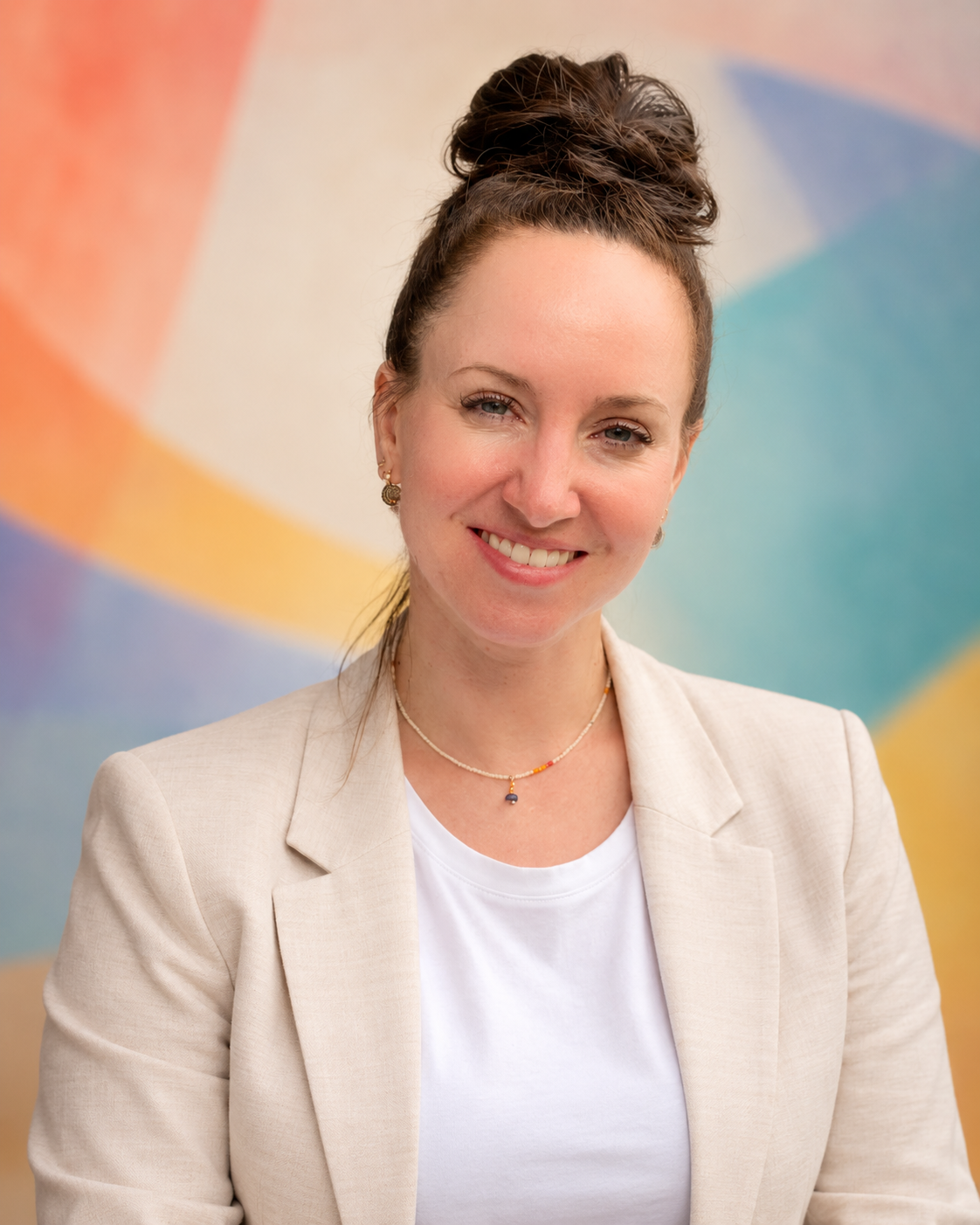 A woman with curly brown hair styled in an updo, wearing a beige blazer and white top, smiling in front of a colorful, abstract background.