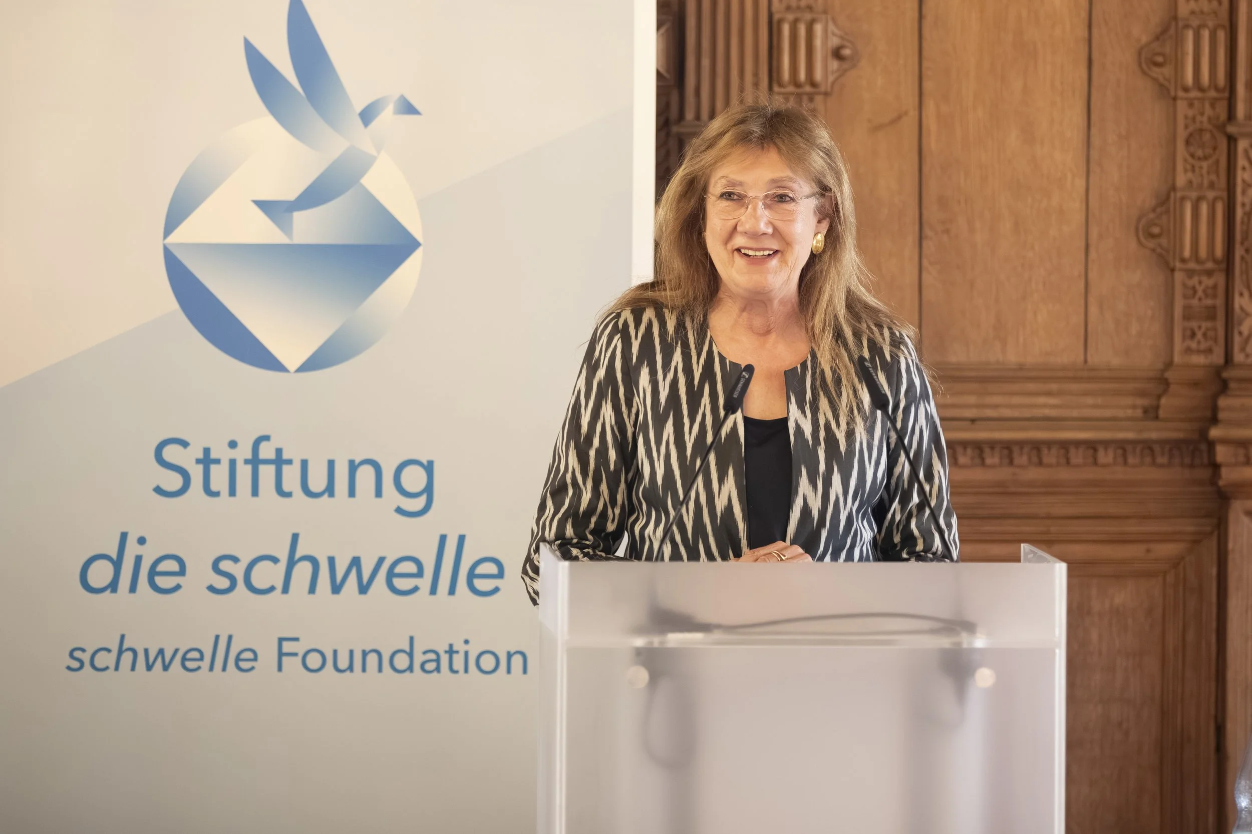 A woman with glasses and shoulder-length hair standing behind a white podium, smiling, with a wooden decorative background. To her left is a large banner with a blue logo and German text that reads 'Stiftung die Schwelle'.