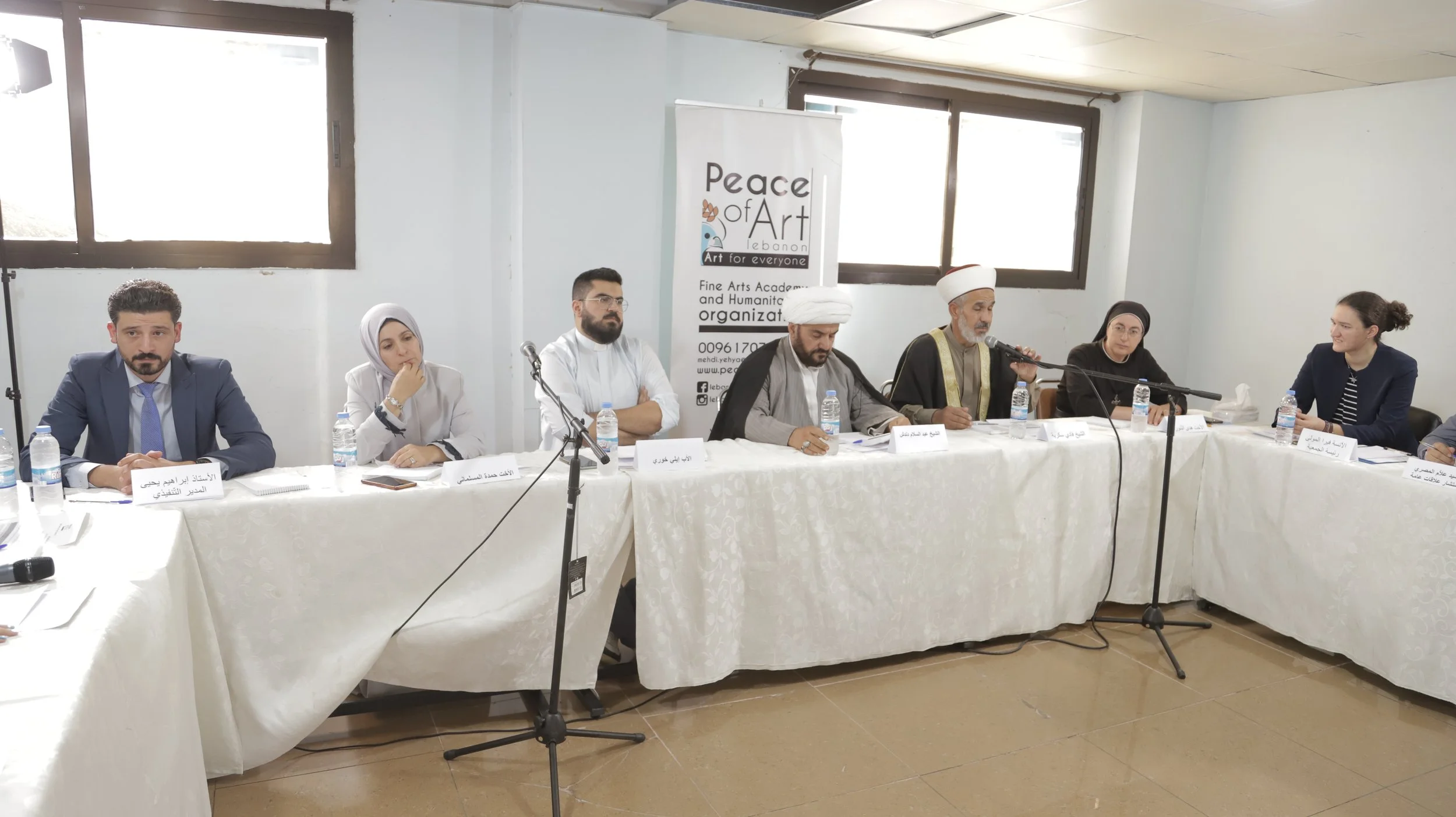 Panel of seven people seated at a long table during a formal meeting or conference, with microphones, water bottles, and nameplates in front of them. Behind them is a banner for Peace of Art Lebanon, an organization related to fine arts and humanitie