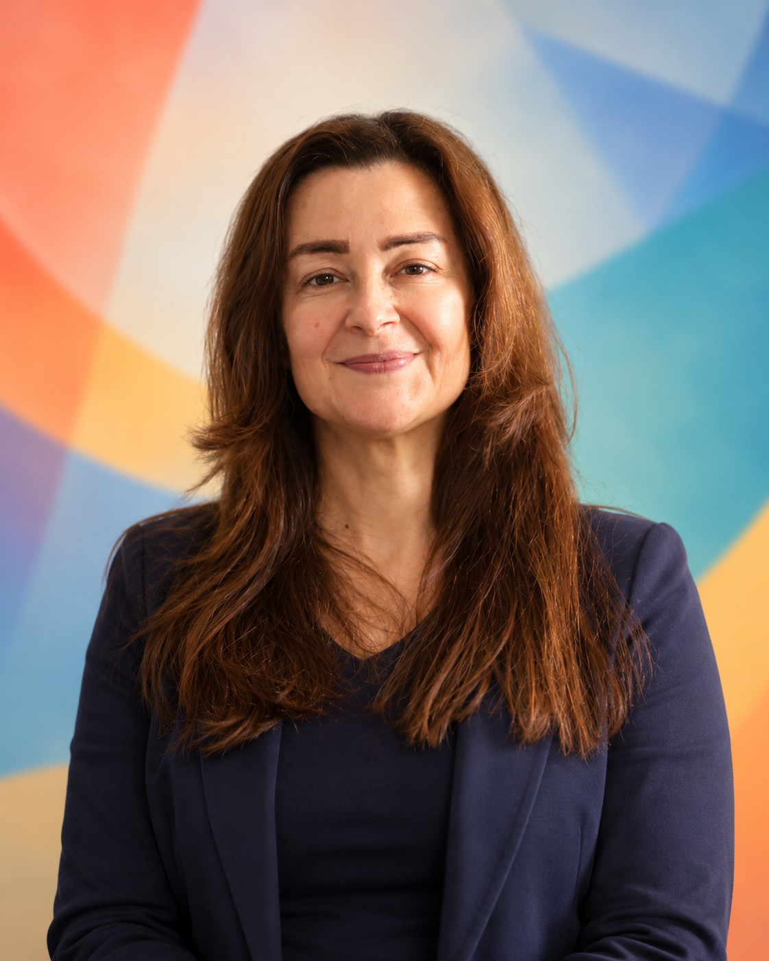 A woman with long brown hair wearing a navy blazer, smiling at the camera, with a colorful geometric background.
