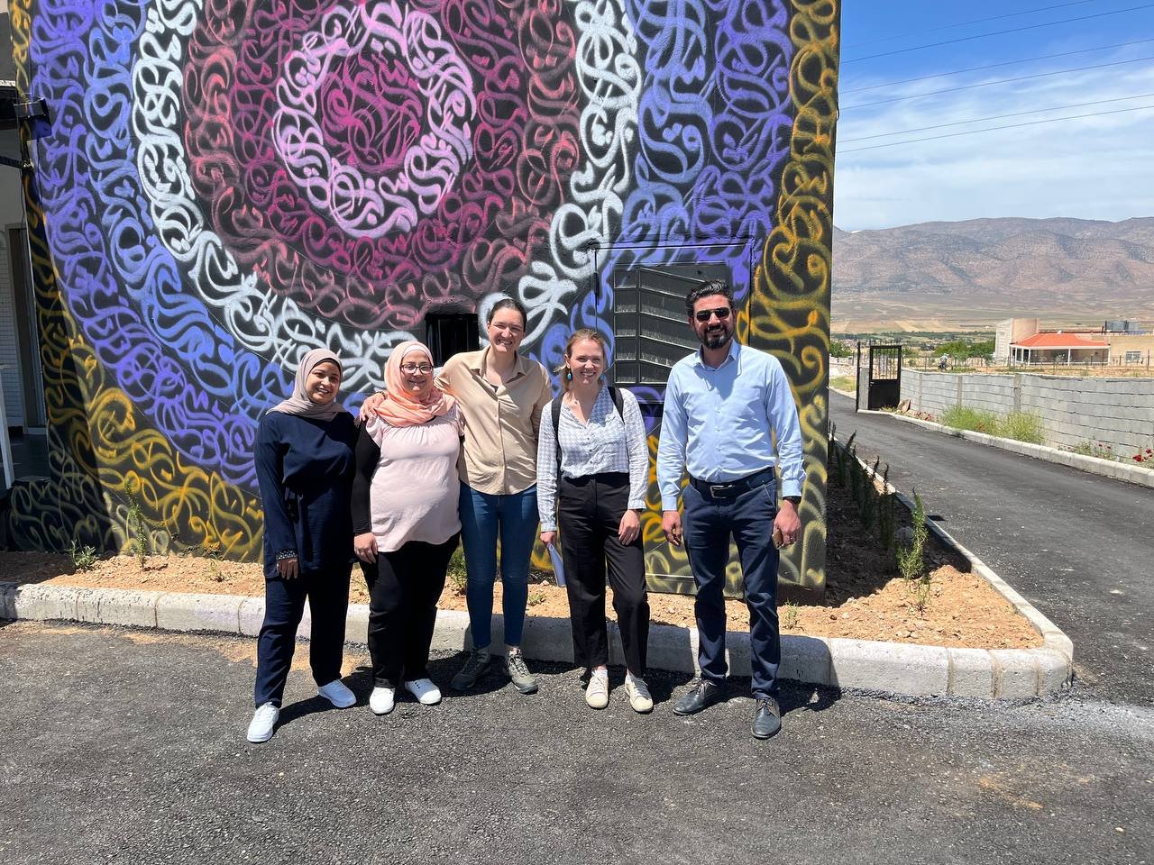 Group of five people standing outdoors in front of a colorful abstract mural on a wall, with mountains and a partly cloudy sky in the background.