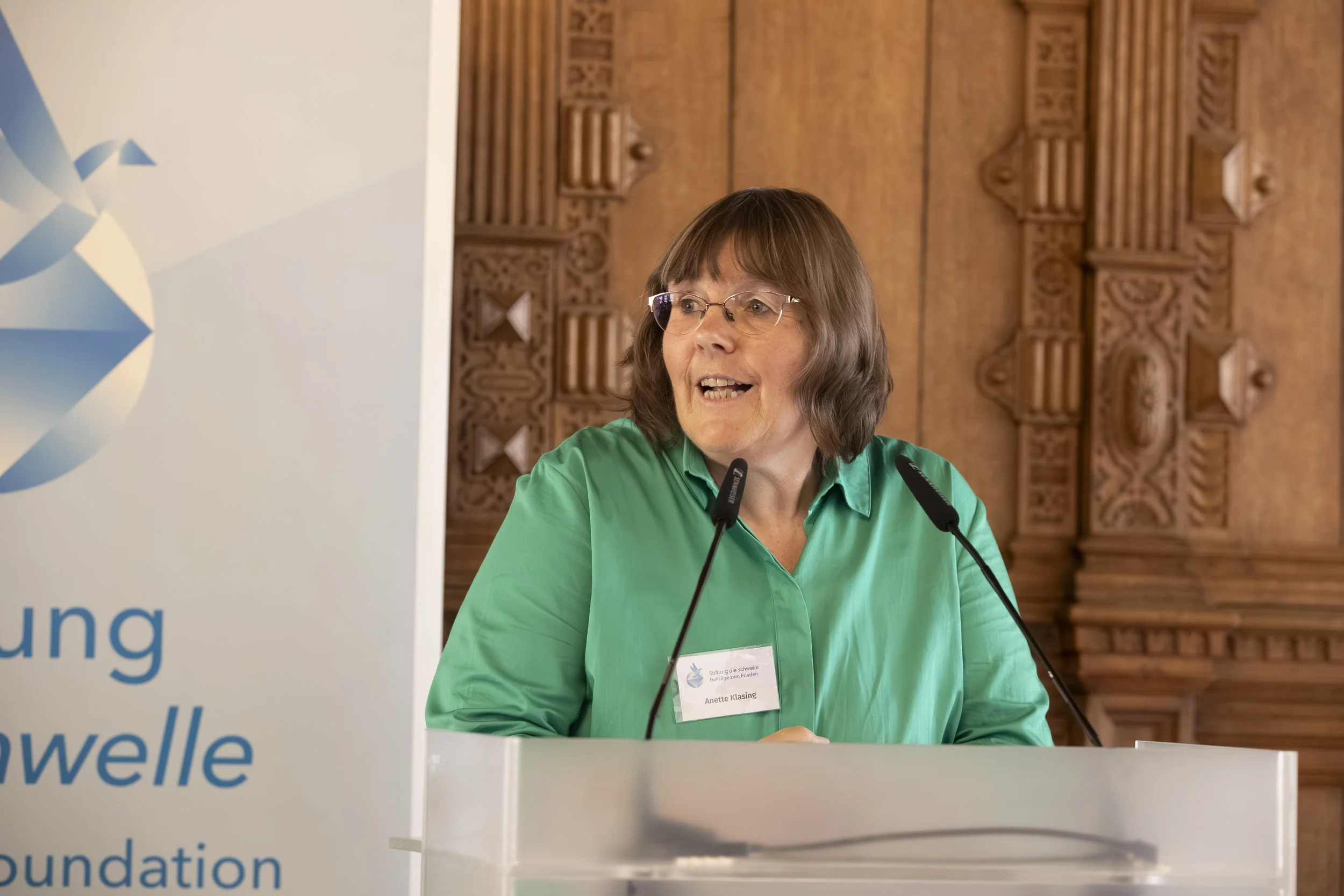 A woman with glasses and shoulder-length brown hair, wearing a green shirt, speaking at a conference with a microphone. She has a name tag that reads 'Anette Klasing.' Behind her is a wooden panel with intricate carvings, and to her left is a white banner with blue text.