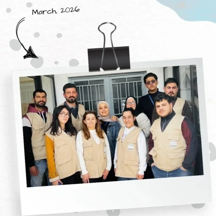 A group of nine people wearing beige vests posing for a photo in front of a glass door. The photo is labeled 'March, 2026' with a drawn arrow pointing to the picture, and a large paperclip graphic is above the photo.