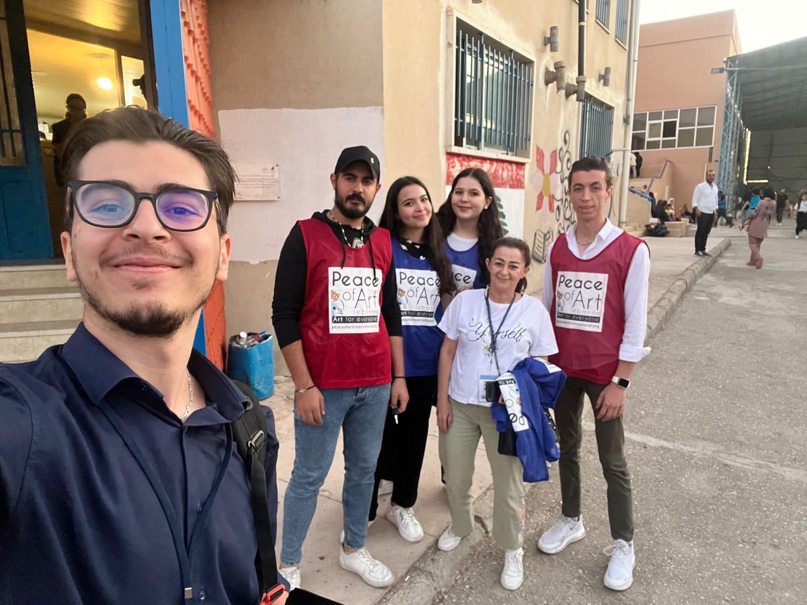 Group of five young people standing outside on a sidewalk during the daytime. Four are wearing red and blue vests with 