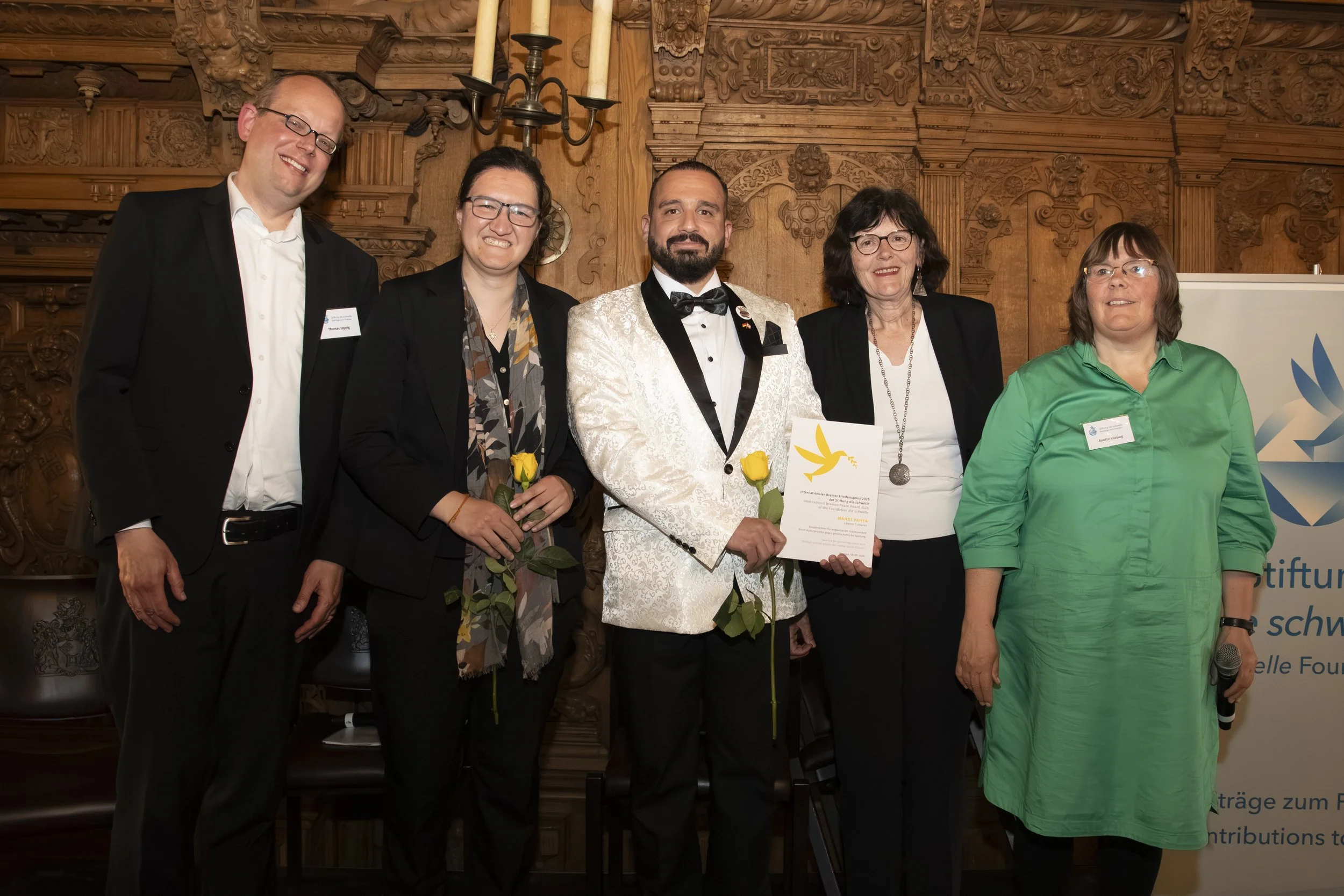 Group of five people standing together indoors, dressed in formal and semi-formal attire, smiling for the camera. The background features elaborately carved wooden decor.