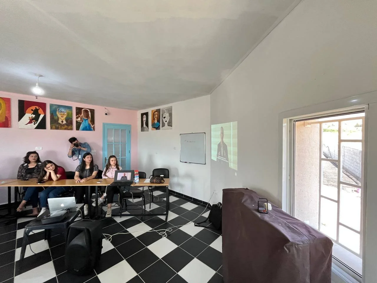 A small conference room with five women, a projector, and a whiteboard. The participants are seated at a rectangular table. Art portraits decorate the wall, and a glass door leads outside.