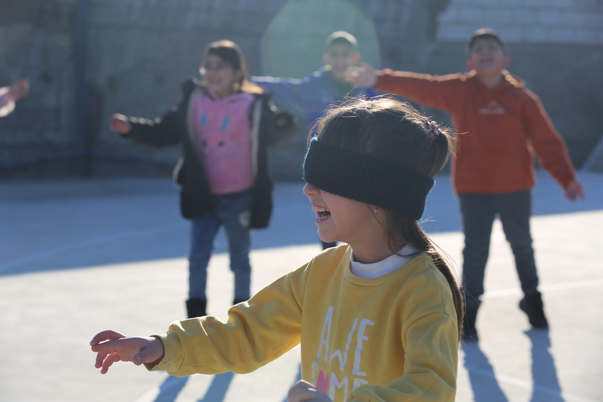 Children playing outside in the snow, with one girl in the foreground wearing a blindfold and a yellow sweatshirt, smiling.