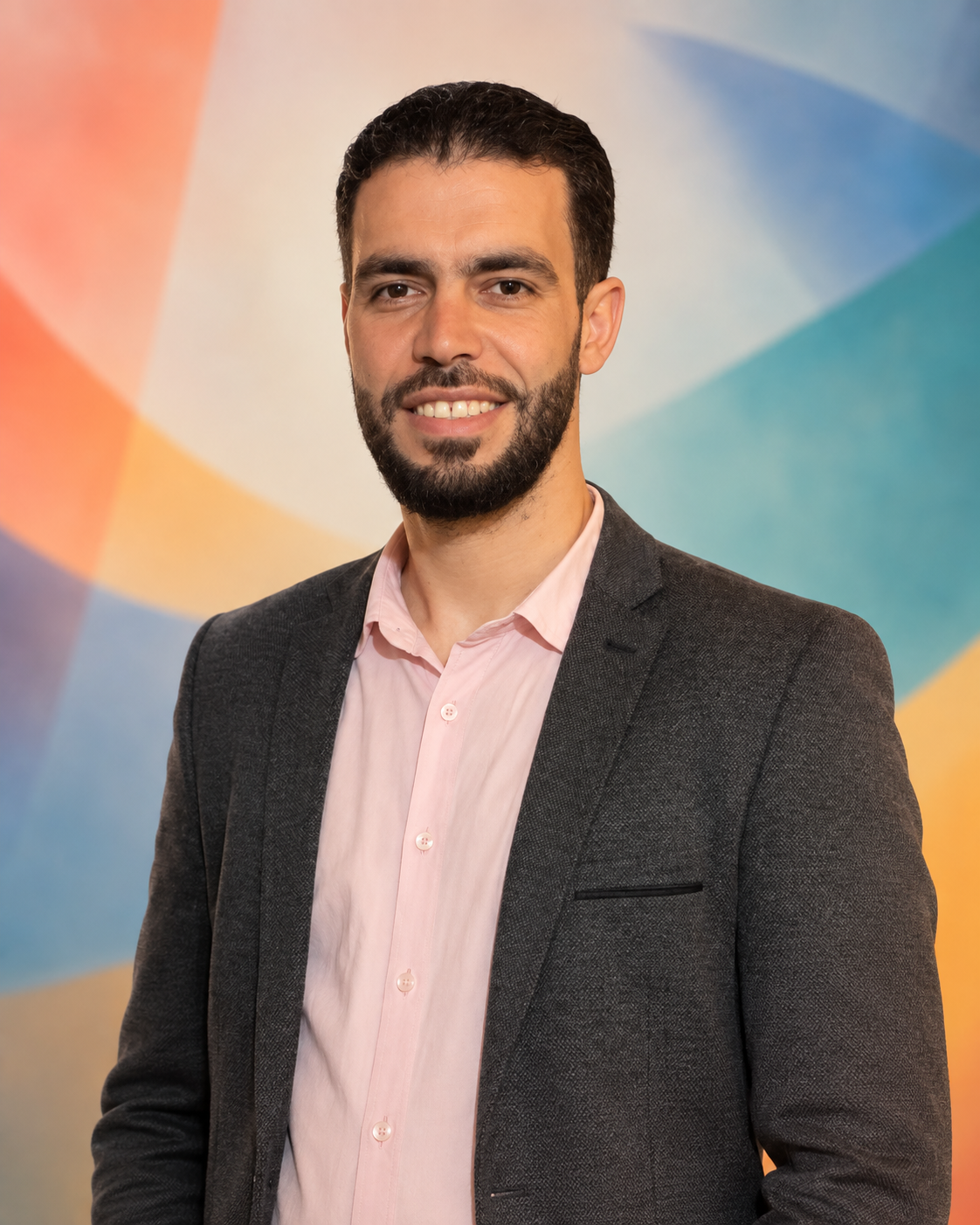 A man with dark hair and beard wearing a dark suit jacket and light pink dress shirt, smiling for a portrait against a design background with a light blue circle and text reading 'NTUR ALDEEN AUSTIN'.