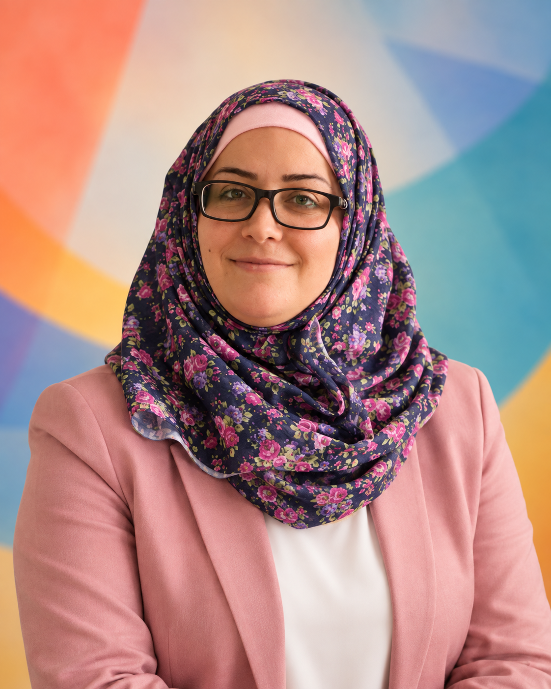 A woman wearing glasses and a floral hijab, dressed in a pink blazer and white top, standing in front of a colorful, abstract background.