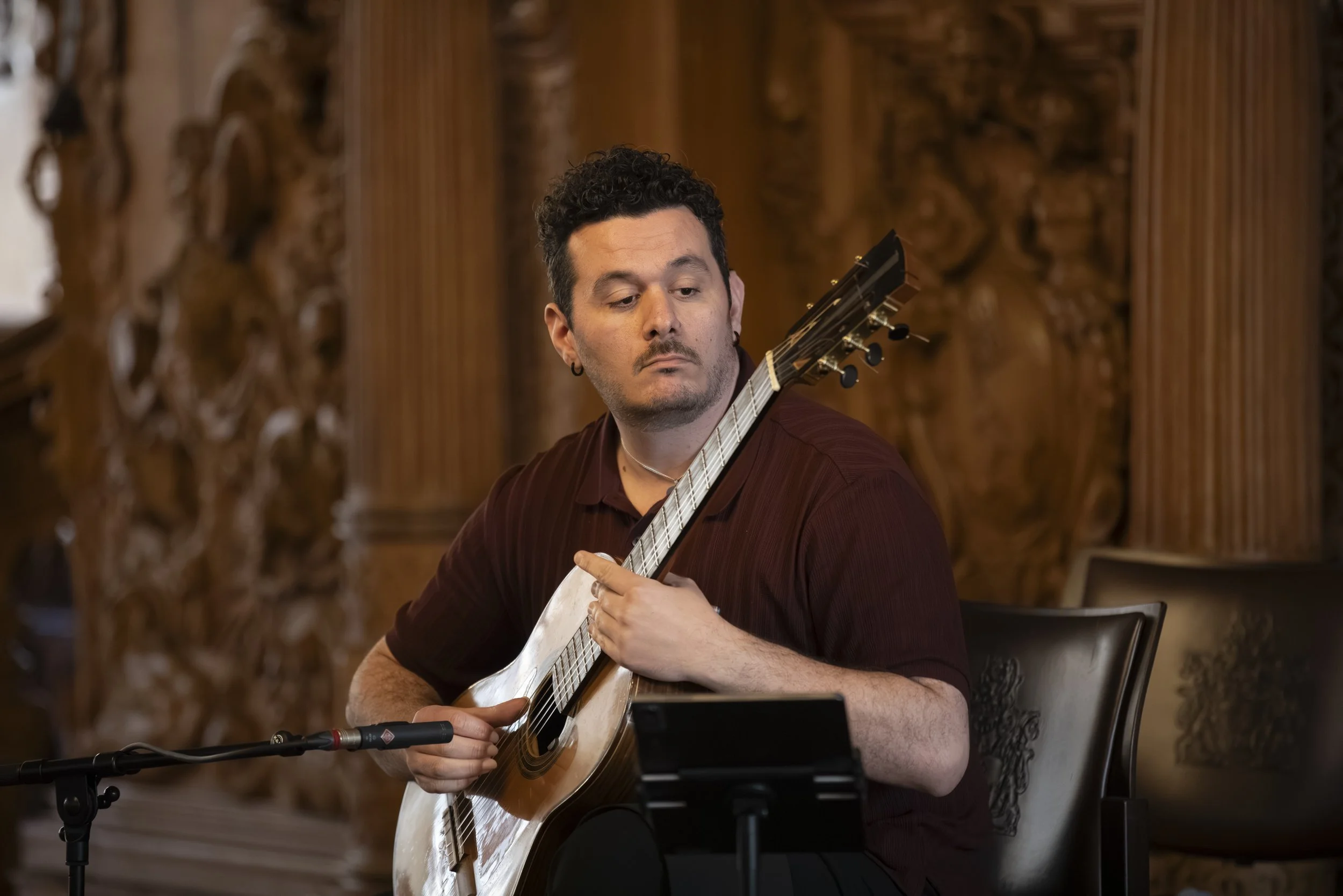A man with dark, curly hair and a small earring is playing an acoustic guitar in a warmly lit room with ornate wooden wall decor. He is wearing a maroon shirt and appears focused on his playing.