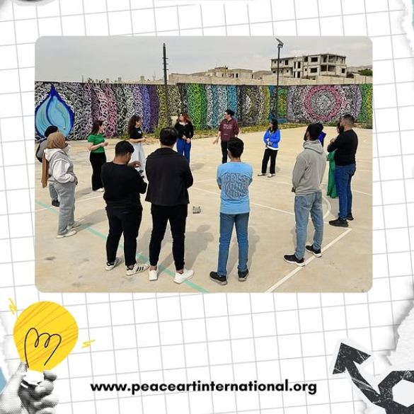Group of people standing in a circle in an outdoor basketball court, with a colorful mural wall and some buildings in the background.