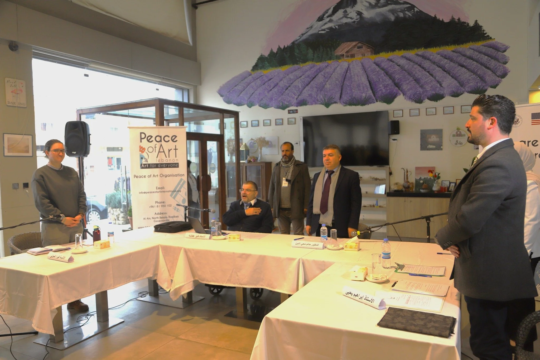 Group of people attending a meeting or conference in a room with a mountain and lavender mural on the wall. Some are seated, and others are standing, with a banner that reads 'Peace of Art' and contact information.