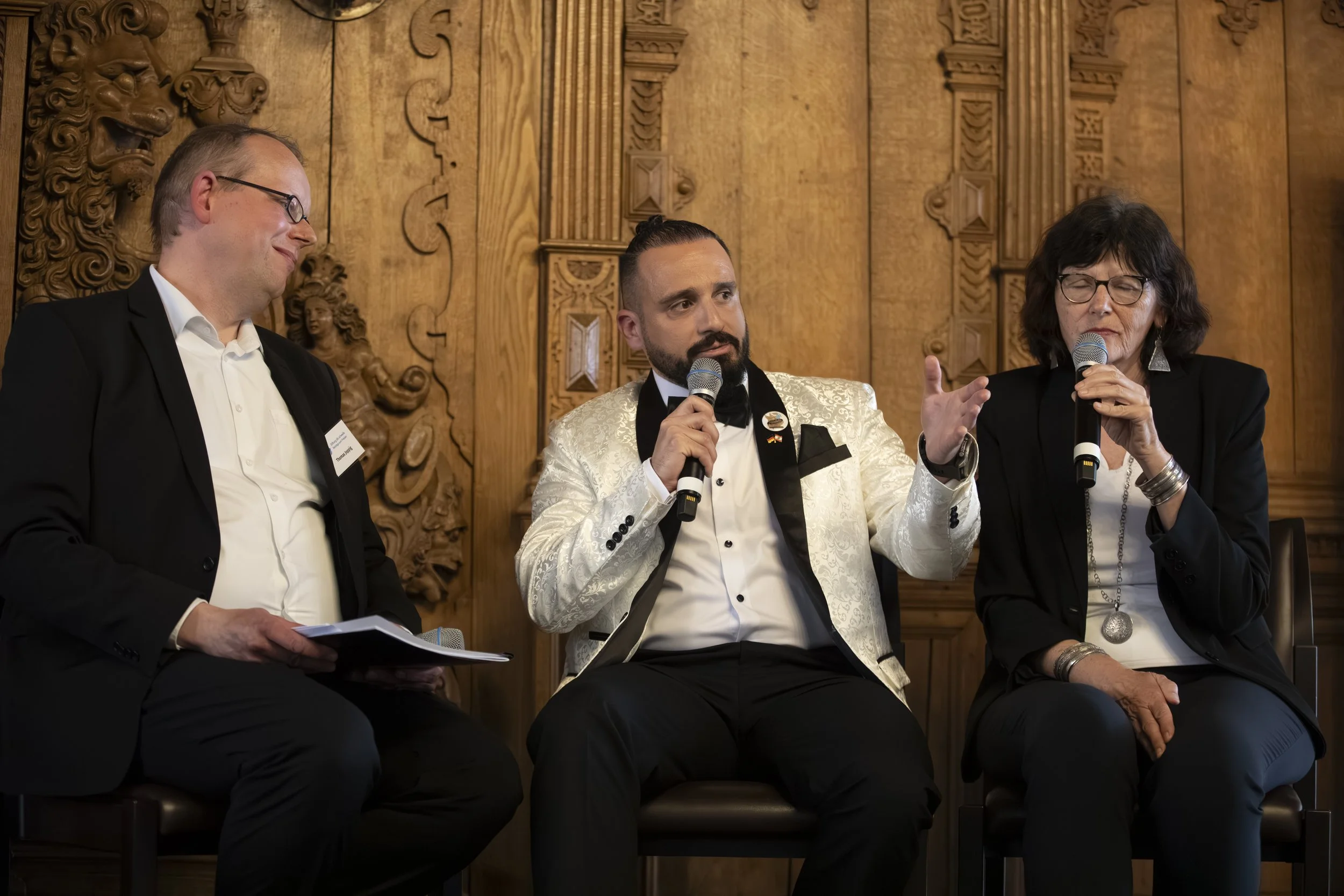 Three people sitting on stage during a panel discussion. The man in the middle is speaking into a microphone, wearing a white suit jacket and black bow tie. The woman on the right is holding a microphone and gesturing with her hand, wearing glasses and a black blazer. The man on the left is looking down at some papers, wearing glasses and a black suit. The background features ornate wood paneling and carved wood details.